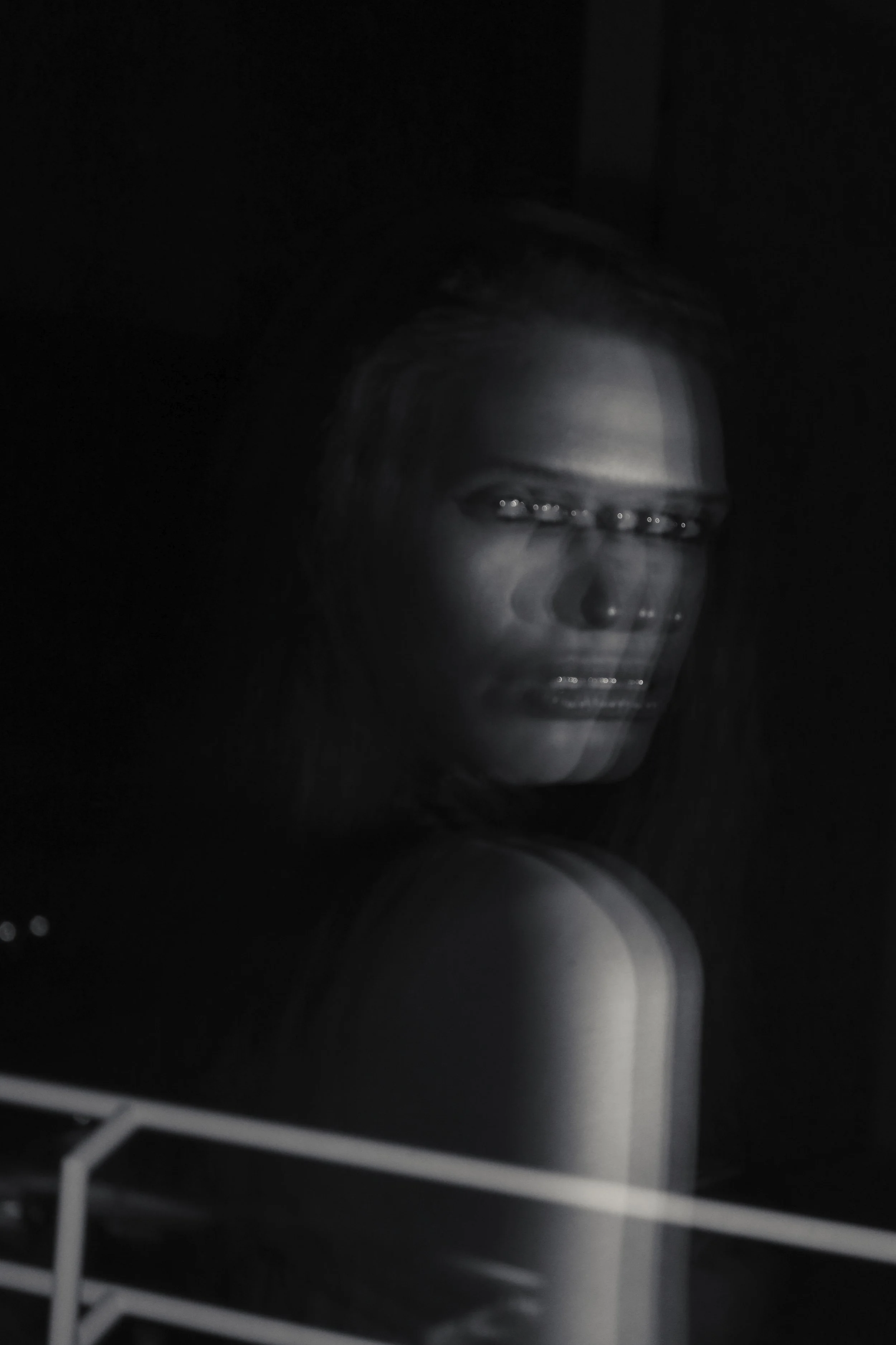 Black and white photograph of a woman viewed through a glass surface, with her face partially obscured by the reflection and the darkness.