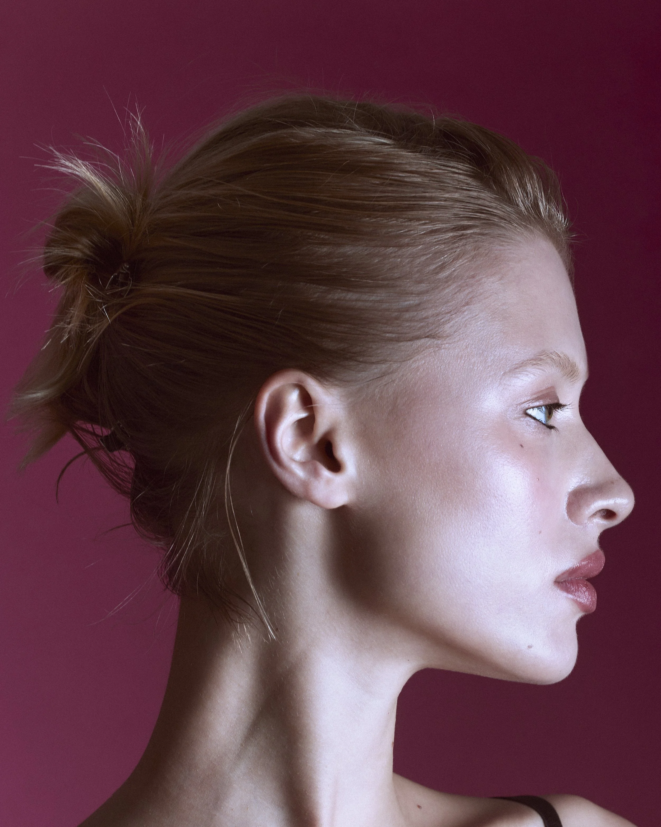 Profile of a woman with fair skin and light brown hair tied back, against a maroon background.