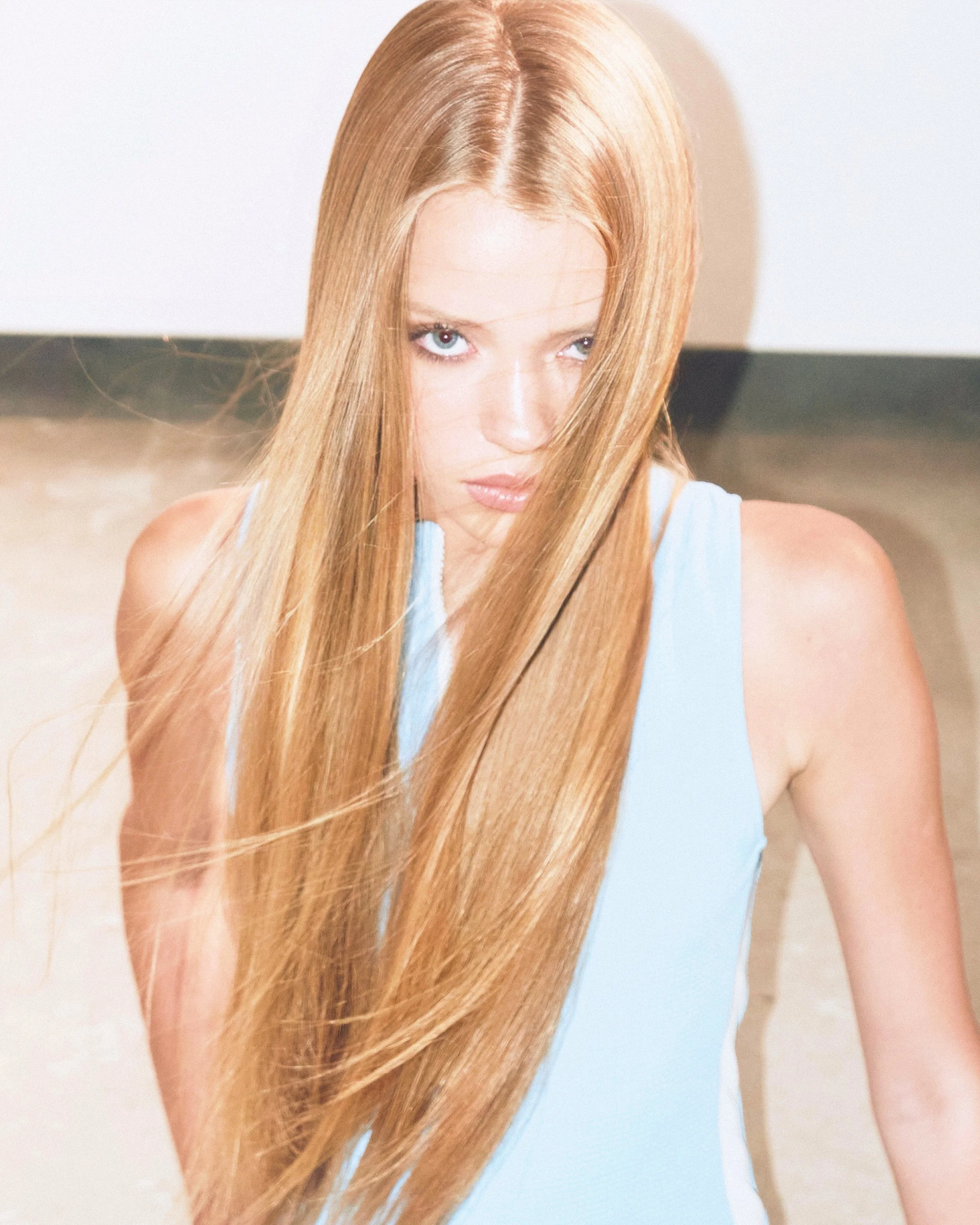 A woman with long, straight red hair and light blue eyes looking upwards at the camera, wearing a light blue sleeveless top.
