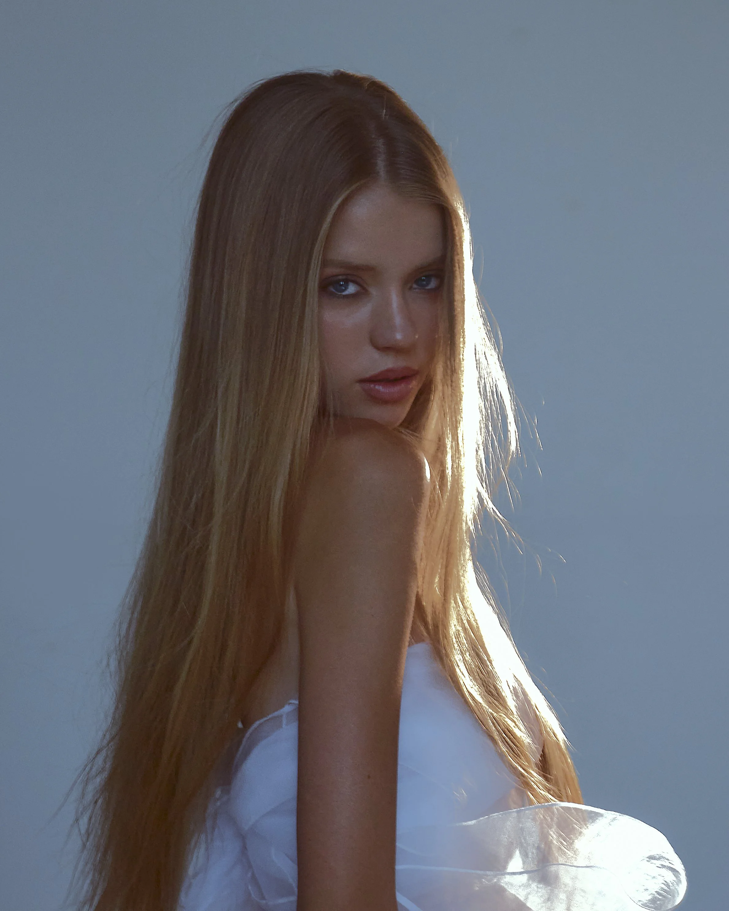 A young woman with long red hair looking over her shoulder, wearing a white top, illuminated by light from behind against a plain gray background.