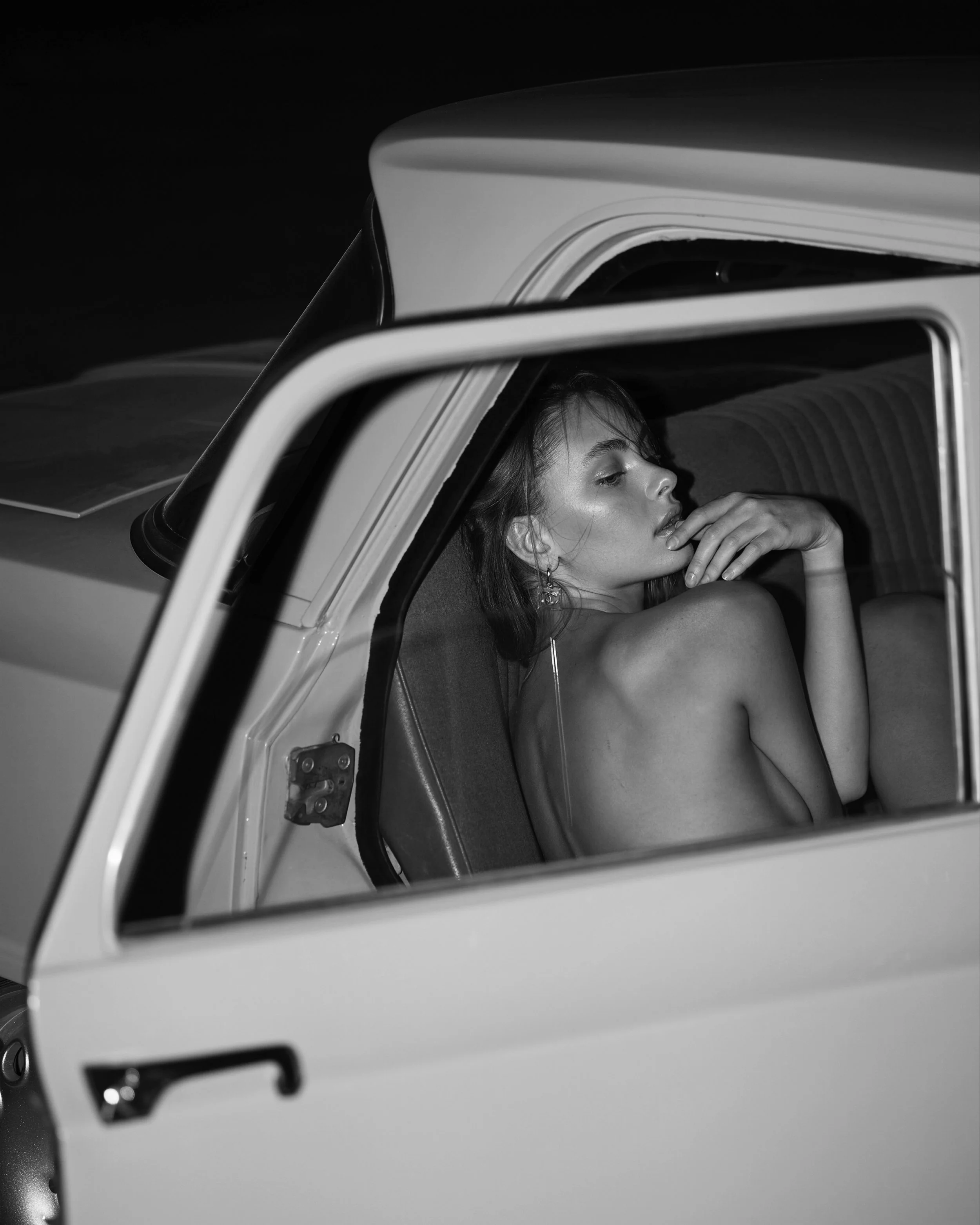 A black and white photo of a woman sitting in the backseat of a car, looking thoughtful with her hand near her face.