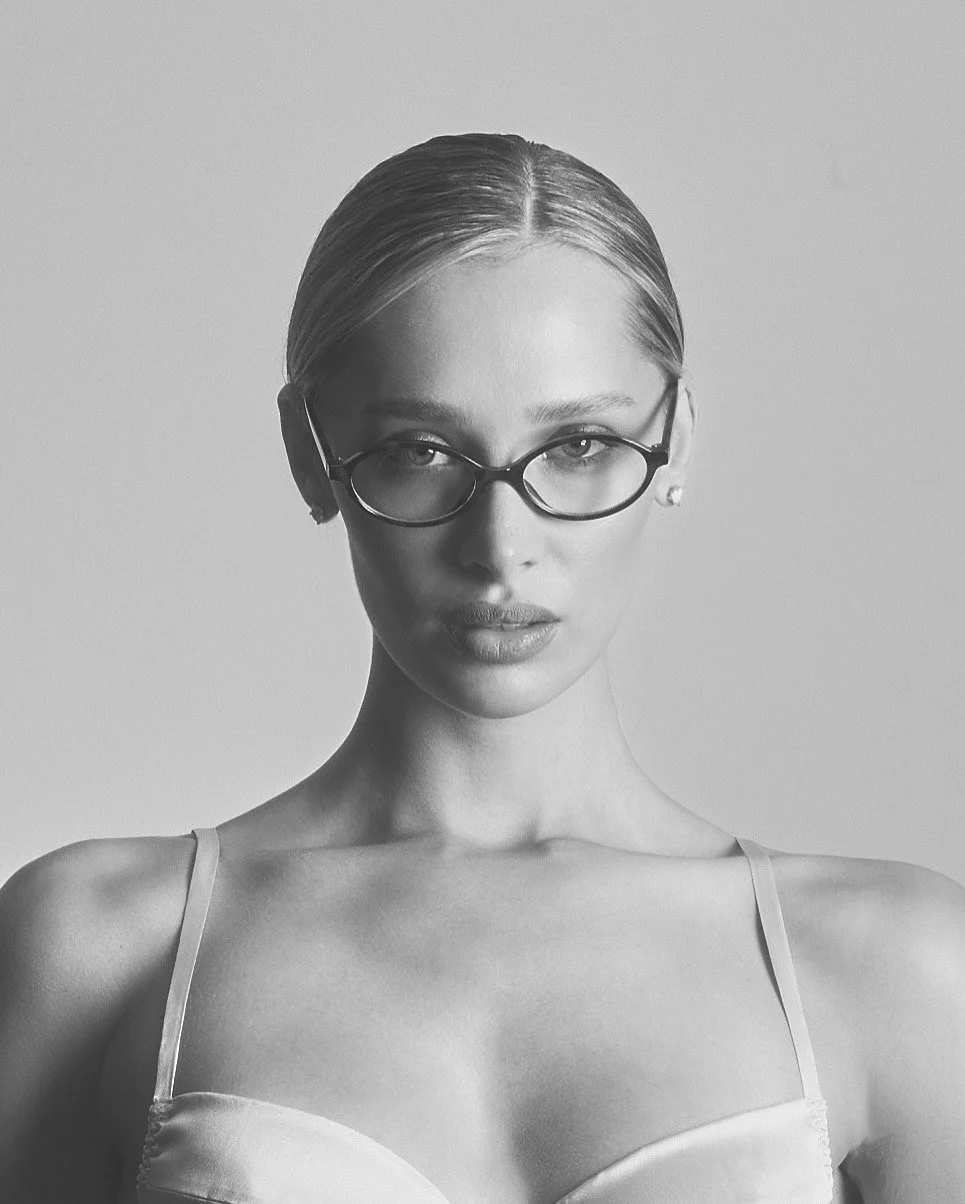 Black and white portrait of a woman with glasses, slicked-back hair, and a spaghetti strap top.
