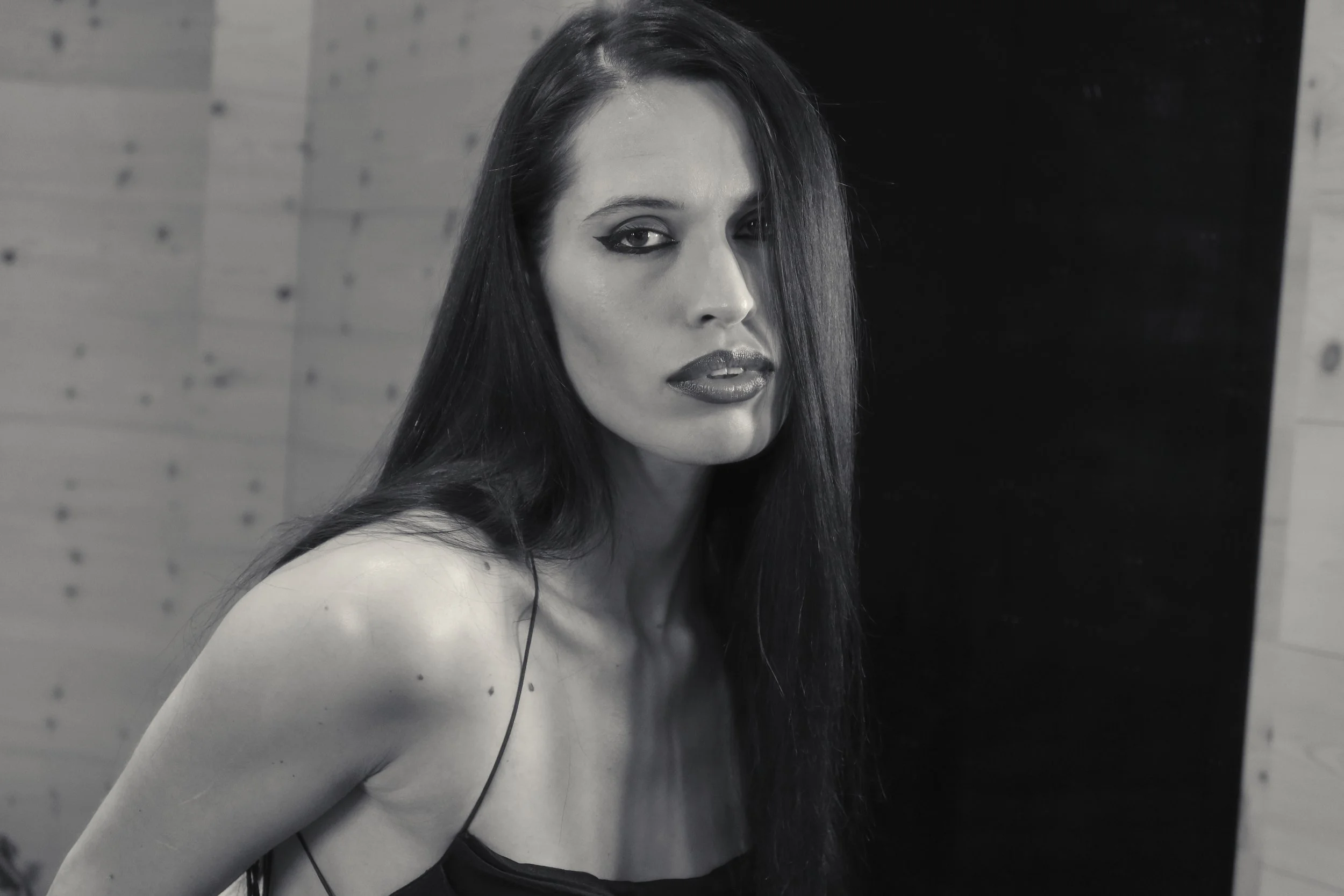 A woman with long dark hair, intense makeup, and a black spaghetti strap top, looking at the camera with a neutral expression in a black and white photograph.