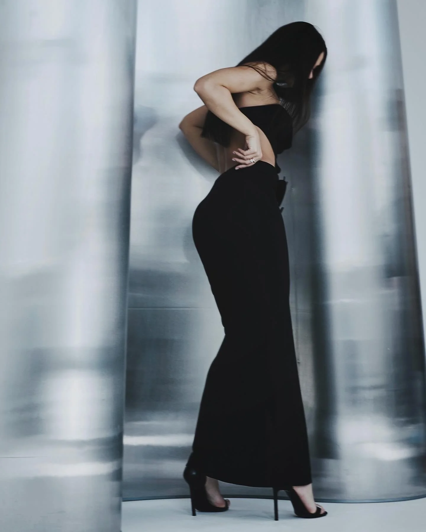 A woman wearing black high heels, black pants, and a black top is standing in an elevator with brushed metal walls, holding a box to her chest.