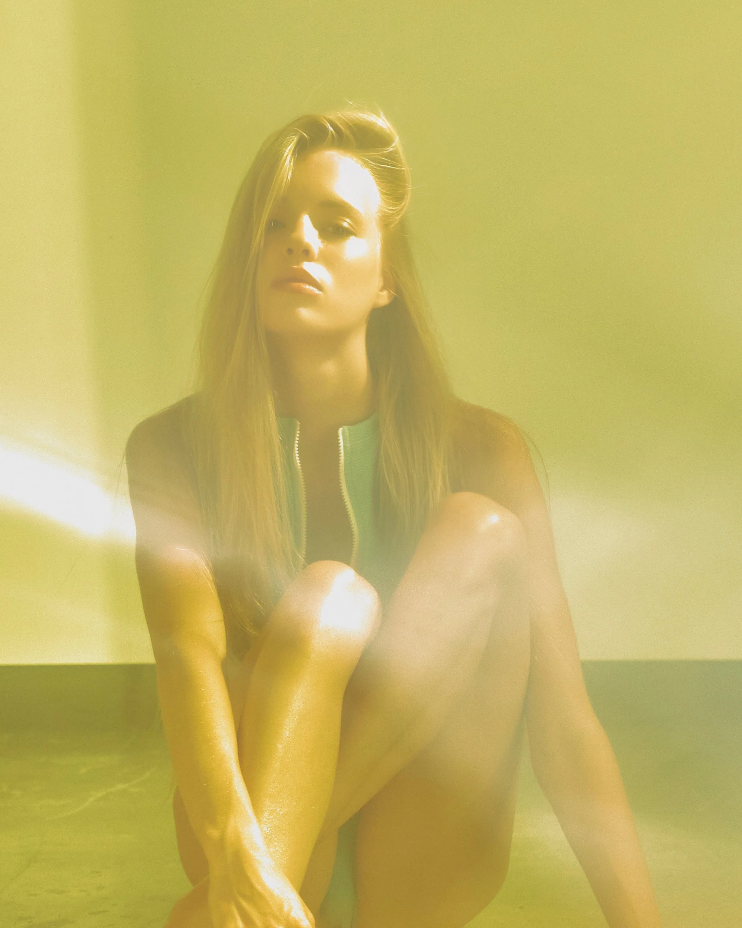 A woman with long blonde hair sitting with her knees drawn to her chest, lit from the side, with a yellowish haze or light in the foreground.