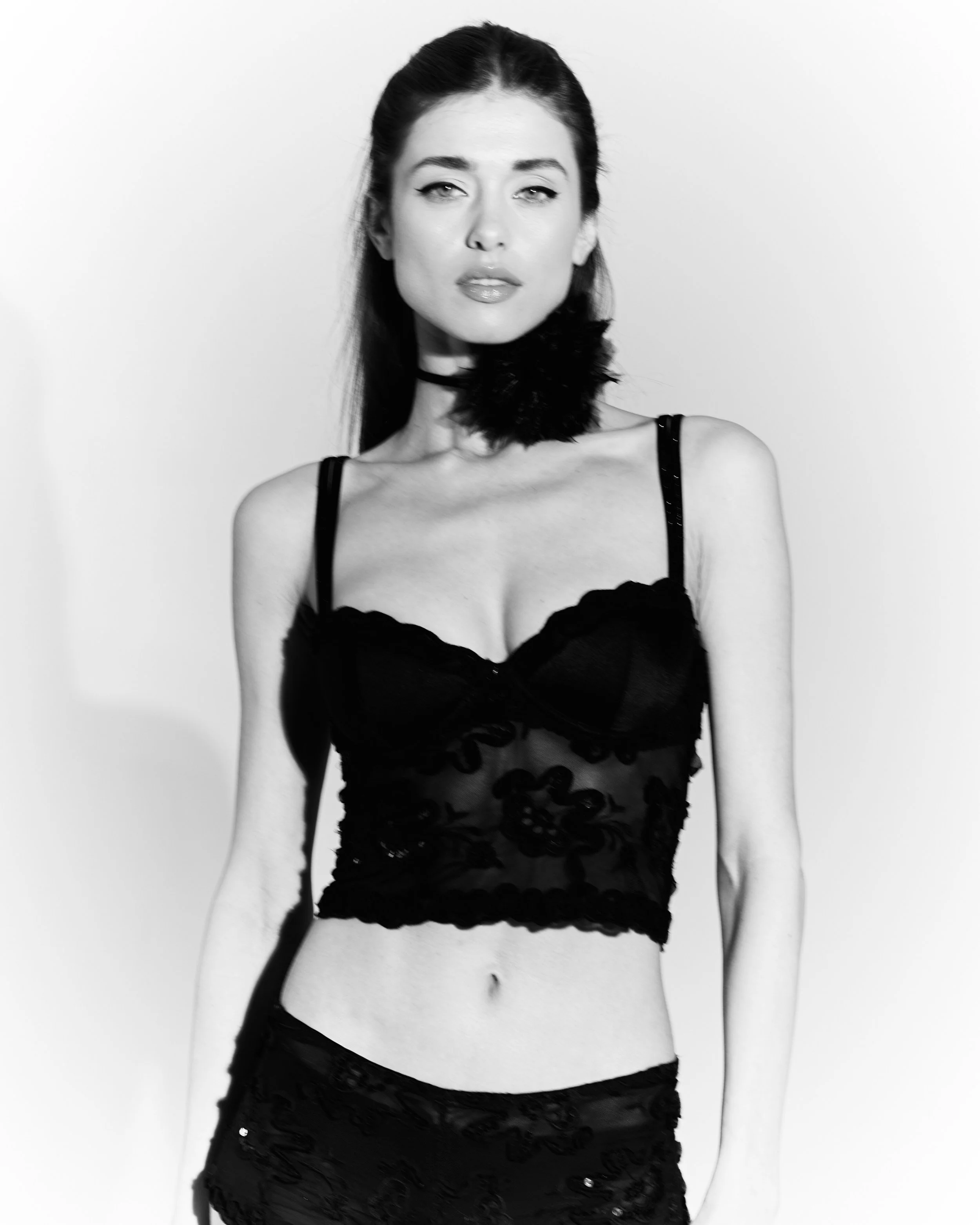 Black and white photo of a woman with dark hair styled straight, wearing a black lace camisole top with floral embroidery and matching short skirt, standing against a plain background.