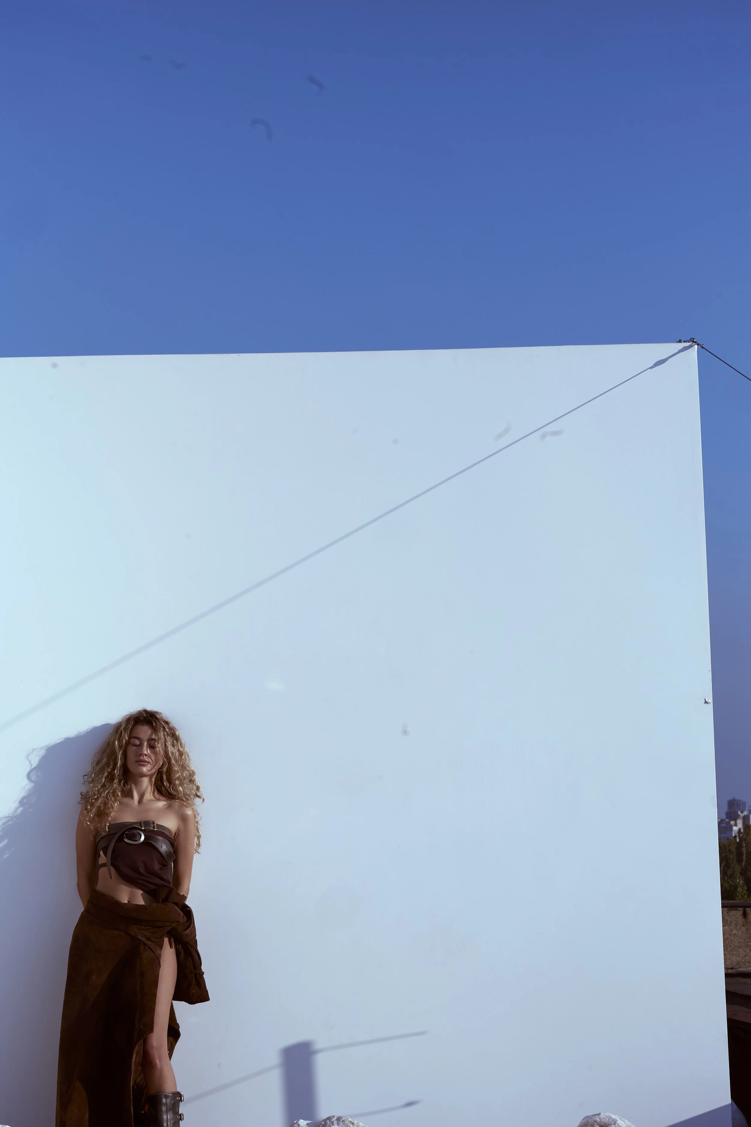 A woman with curly hair standing against a white wall outdoors, with a clear blue sky above.