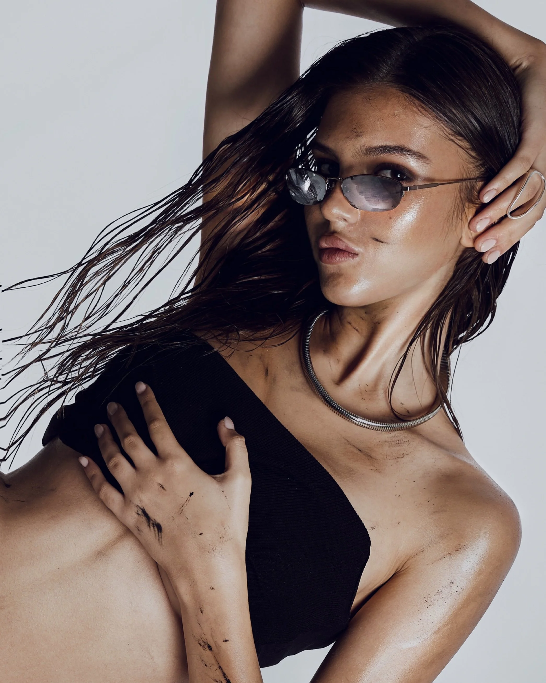 A woman with wet hair wearing sunglasses, a black top, and a silver necklace, posing with one hand on her head and the other on her chest, against a plain background.