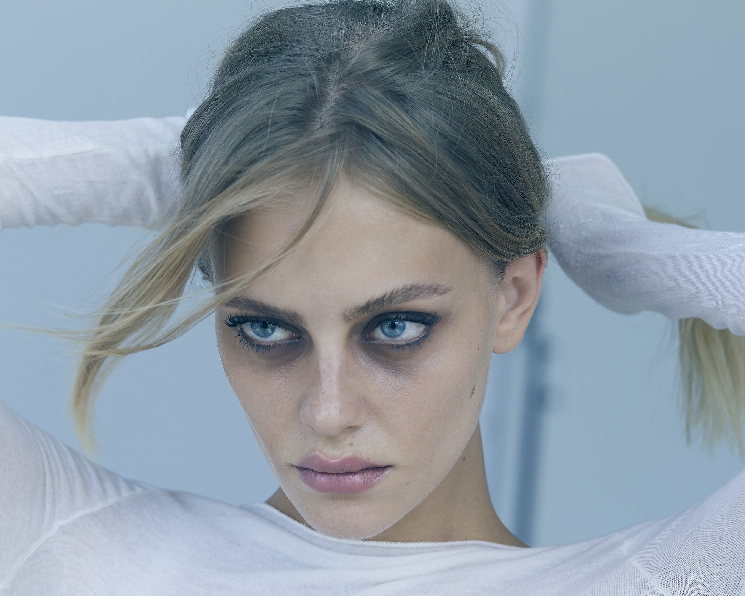 Close-up of a young woman with blonde hair and striking blue eyes, fixing her hair with both hands, wearing a white top.