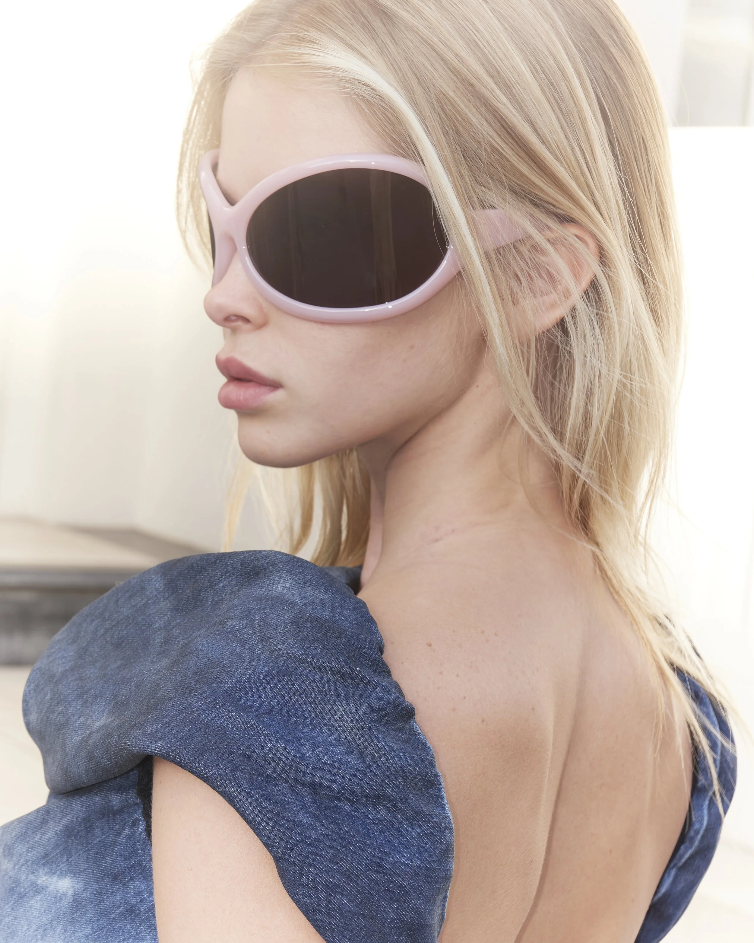 A young woman with blonde hair wearing pink sunglasses and a blue dress with puffed sleeves, looking sideways and off camera.