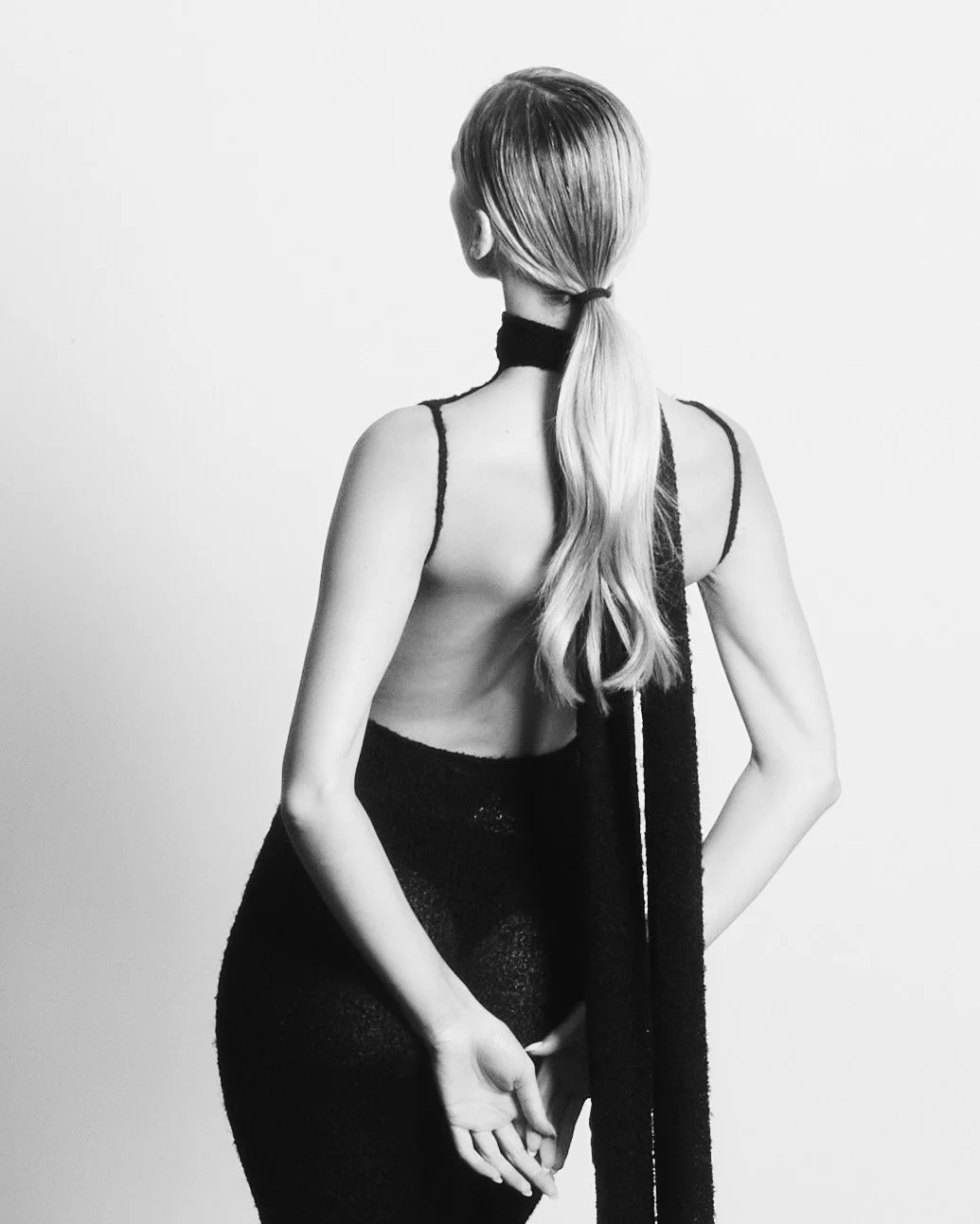 Black and white photo of a woman with long hair in a ponytail, wearing a black dress with thin straps and a black scarf, standing against a plain light background, facing away from the camera.
