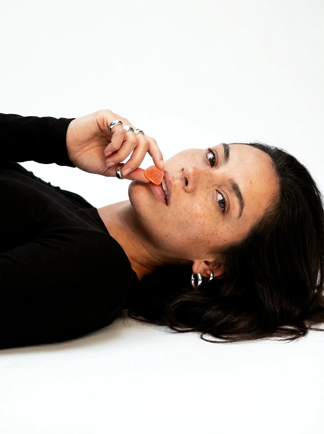 A woman with dark hair and jewelry lying on her side, holding a supplement gummy near her lips, against a plain white background.