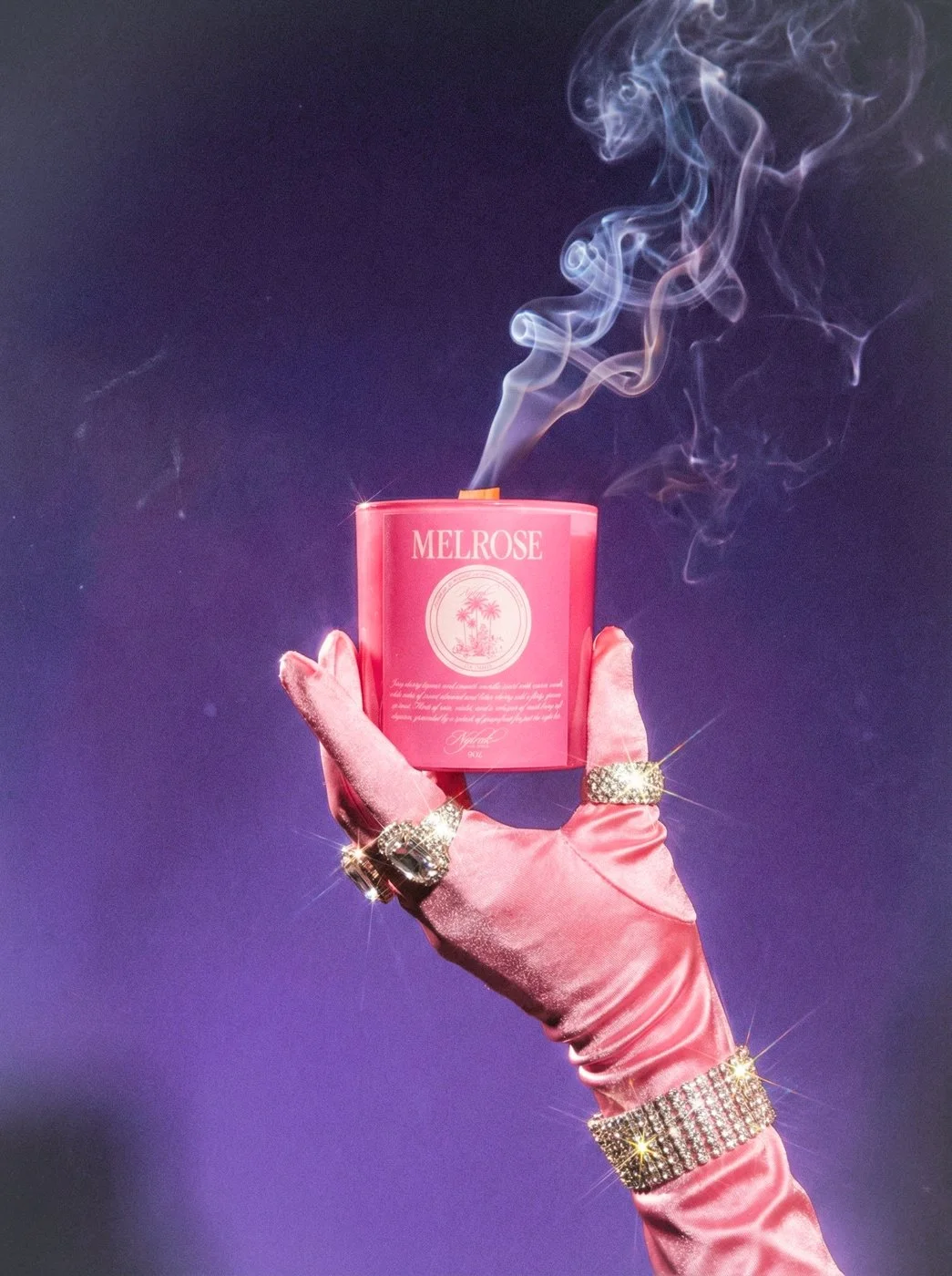 A hand wearing a pink satin glove, adorned with rings and a sparkling bracelet, holds a pink container labeled 'Melrose' with smoke coming out, set against a dark background.