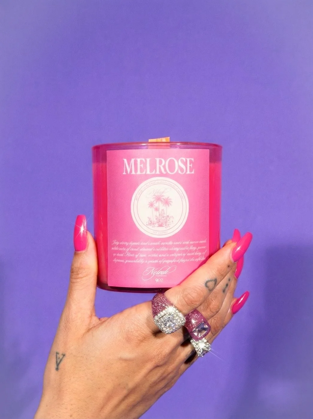 Hand with pink nails and multiple rings holding a pink Melrose scented candle against a purple background.