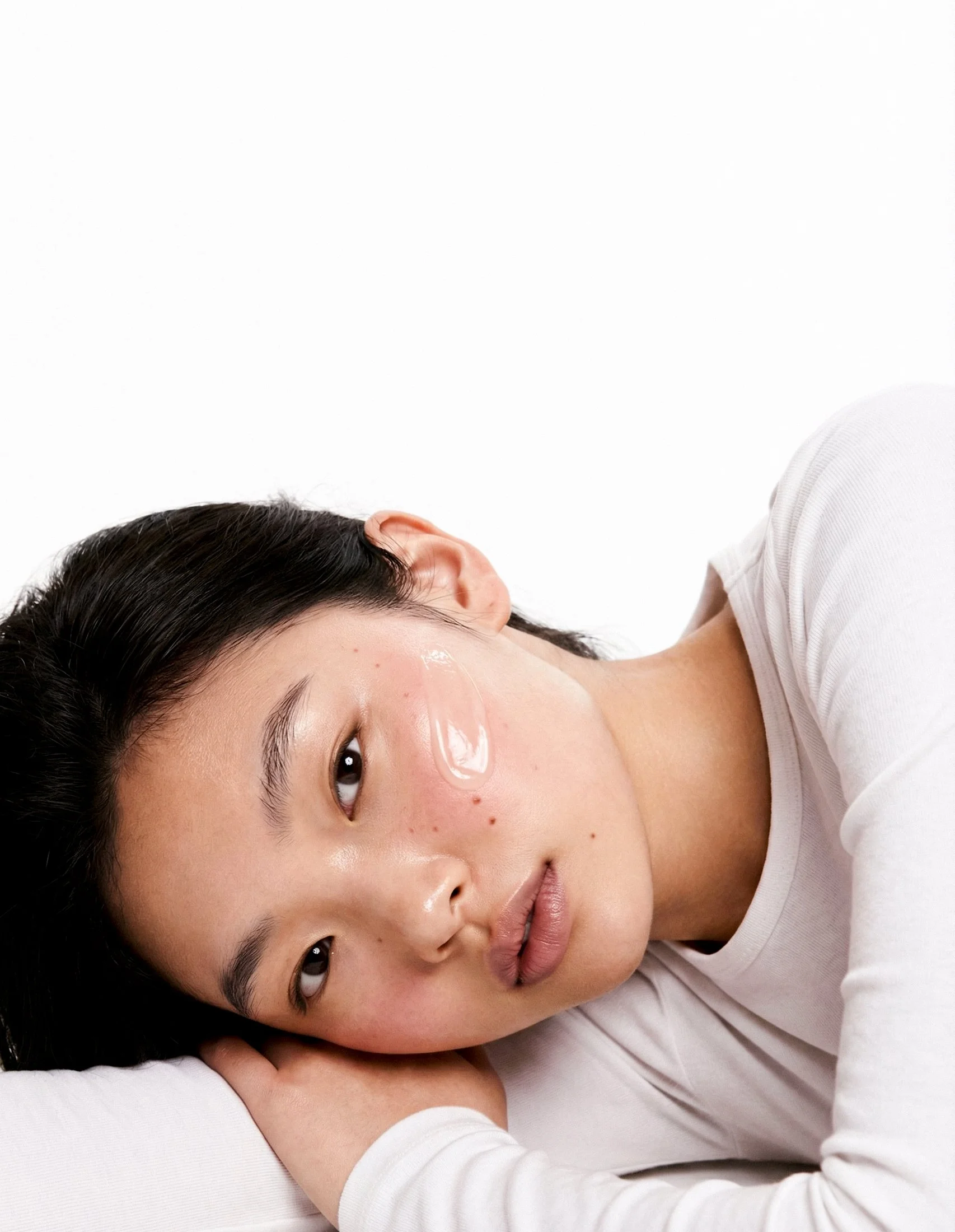 A young woman with black hair lying down on her side, with her head resting on her arm, wearing a white shirt, looking at the camera with a relaxed expression