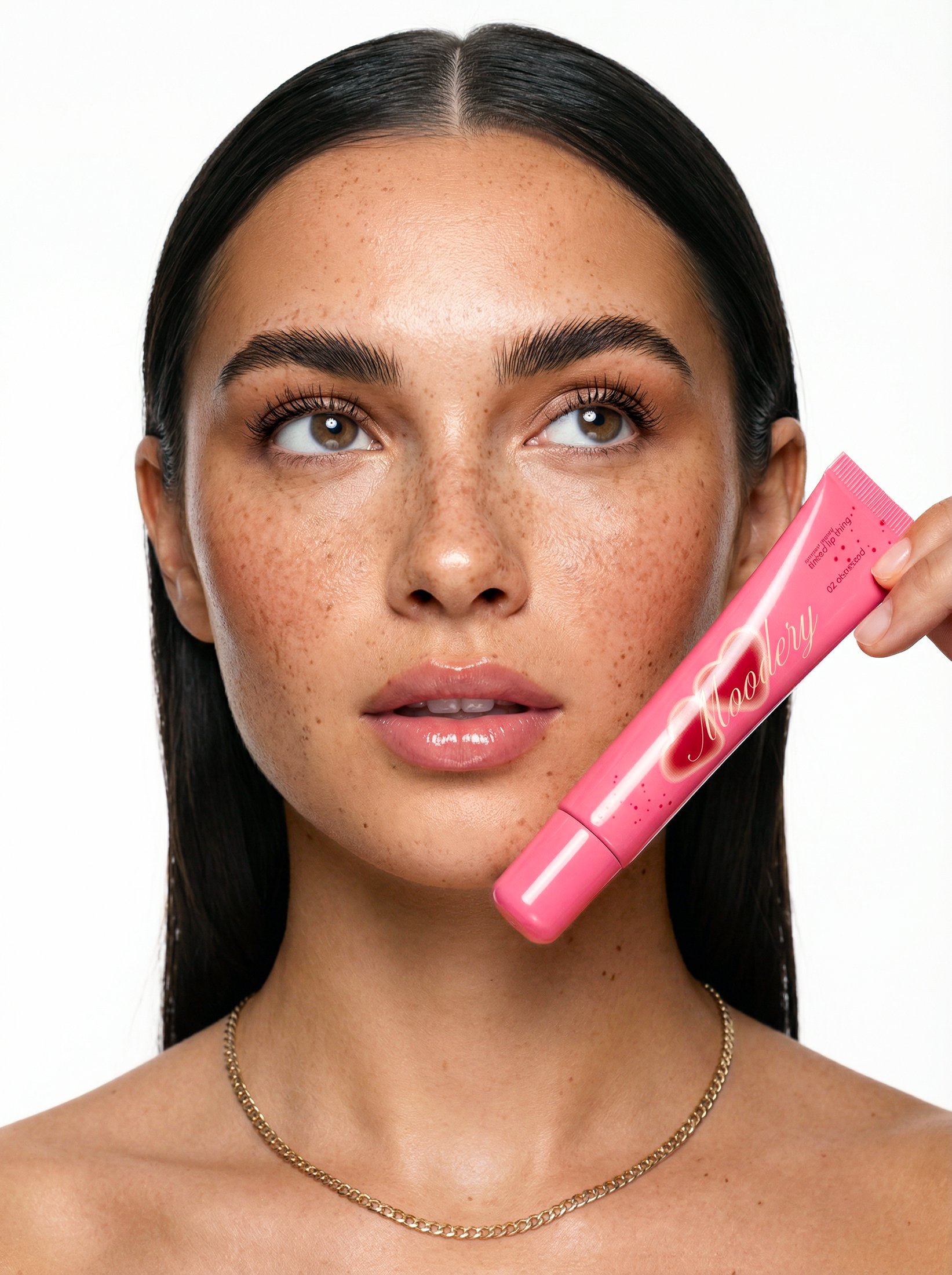 A woman with dark hair, light skin, and freckles holding a pink lipgloss labeled 'Moodery' near her face, against a plain white background.