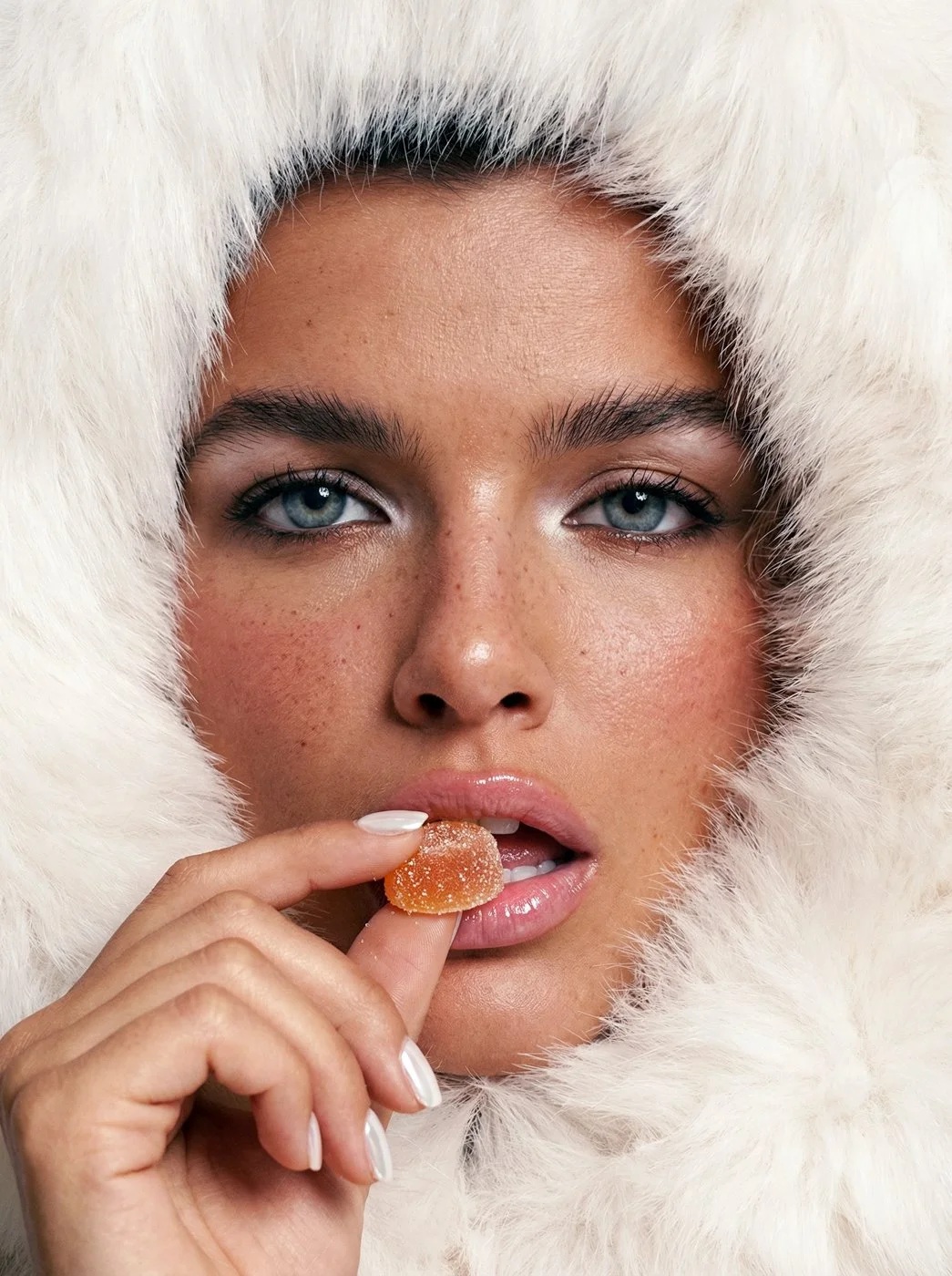 Close-up of a woman with blue eyes and freckles, wearing a white fur hood, holding a gummy supplement near her lips