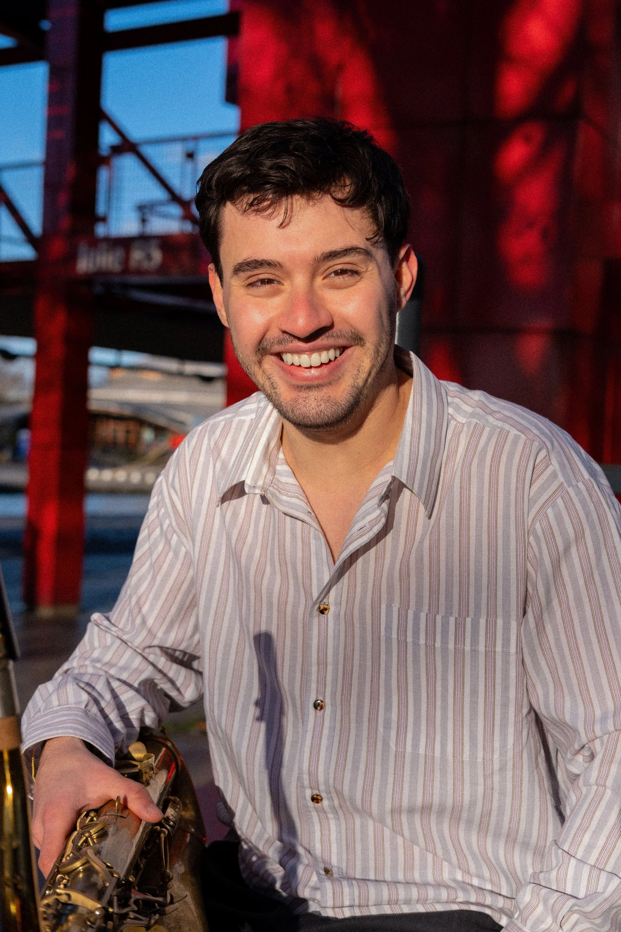 A smiling man with dark hair and a beard, wearing a striped button-up shirt, sitting outdoors near a table with a saxophone, with a red and blue background.
