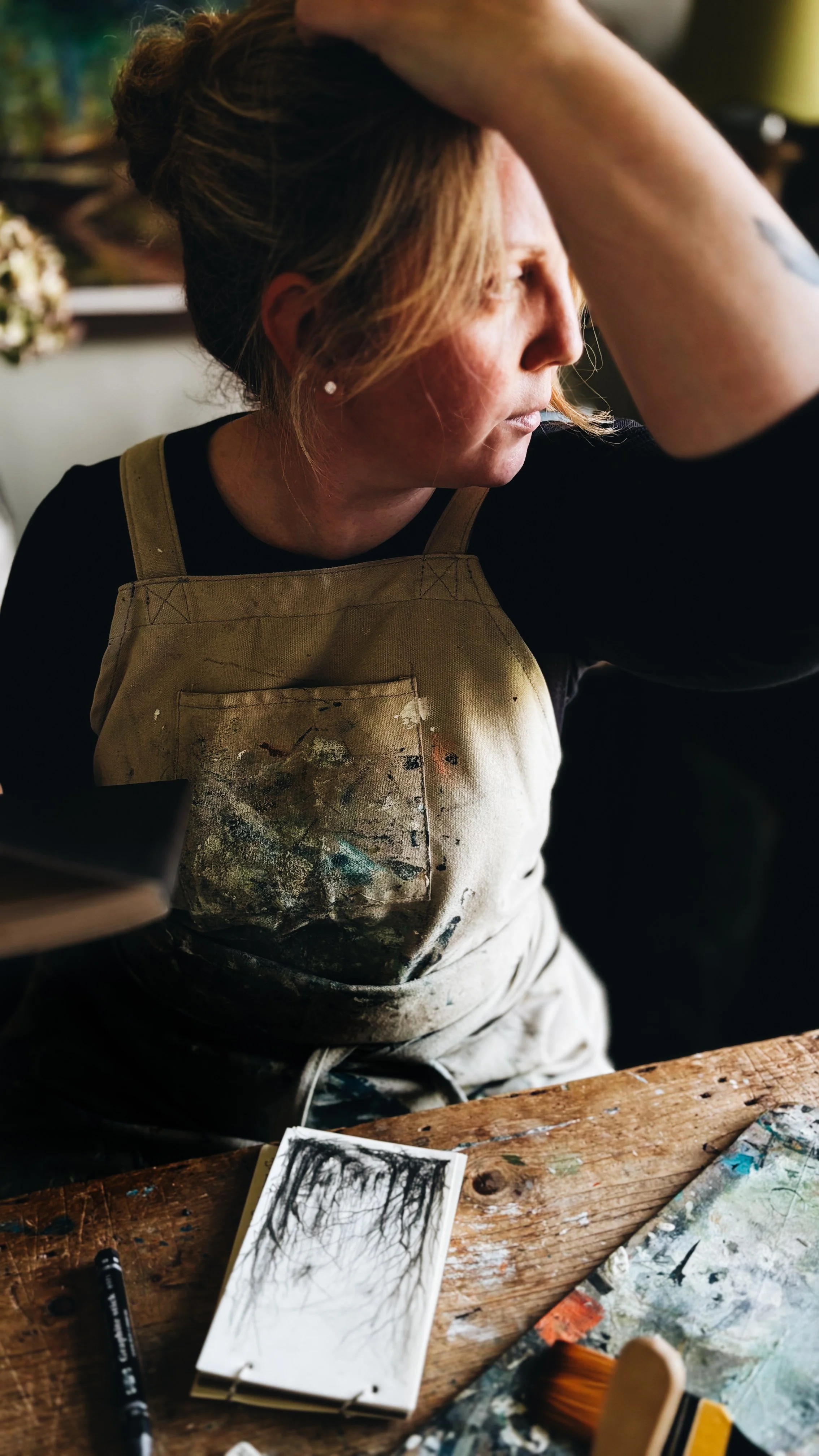 artist Victoria Hodgson of northern art house sits in her studio wearing a painting apron with her hair up in a messy bun. she is looking into the distance with her arm raised as if in contemplation.