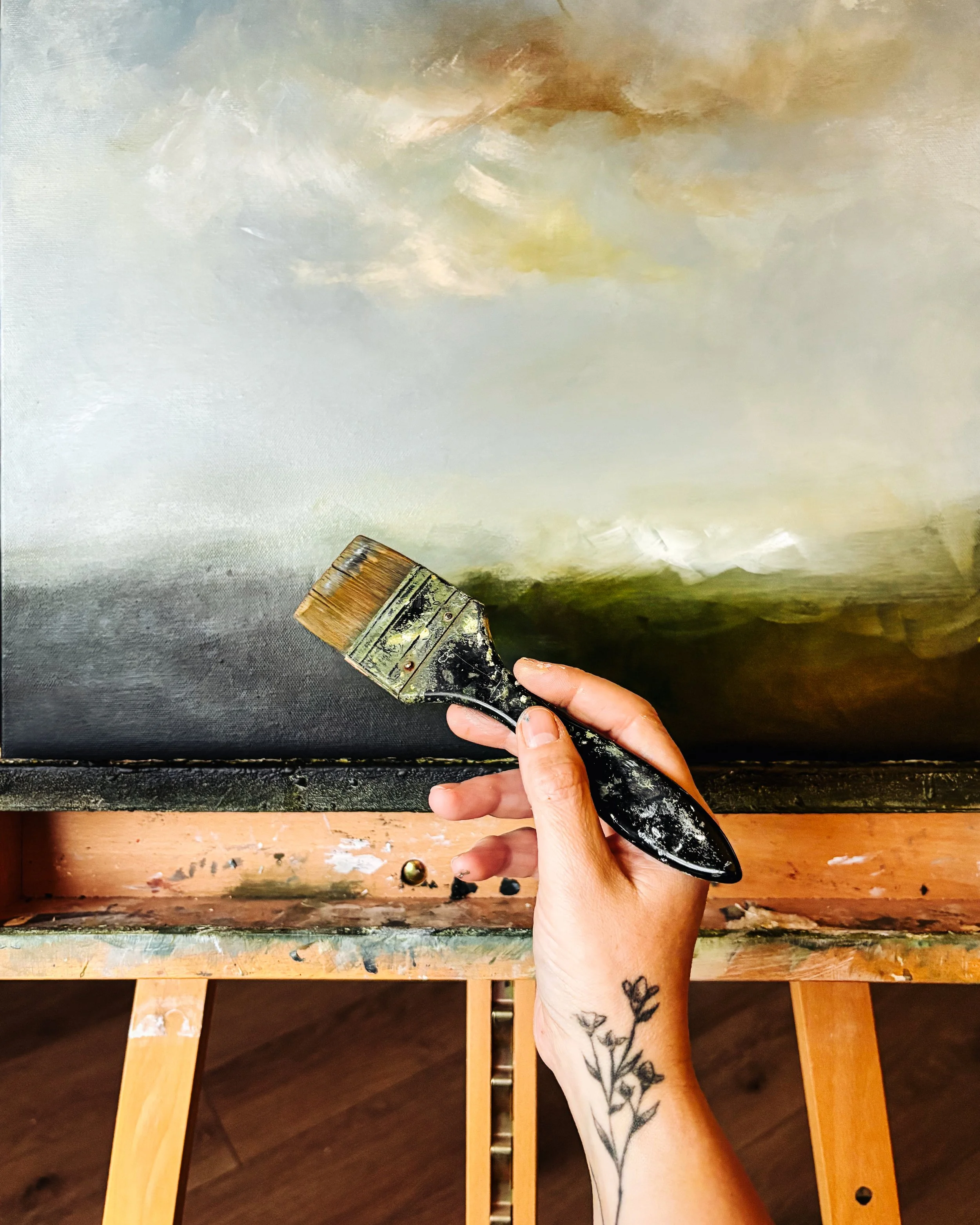 A person's hand holding a paintbrush with black paint, poised above a canvas with an abstract painting in shades of white, brown, yellow, black, and gray, with a wooden table and chair underneath.