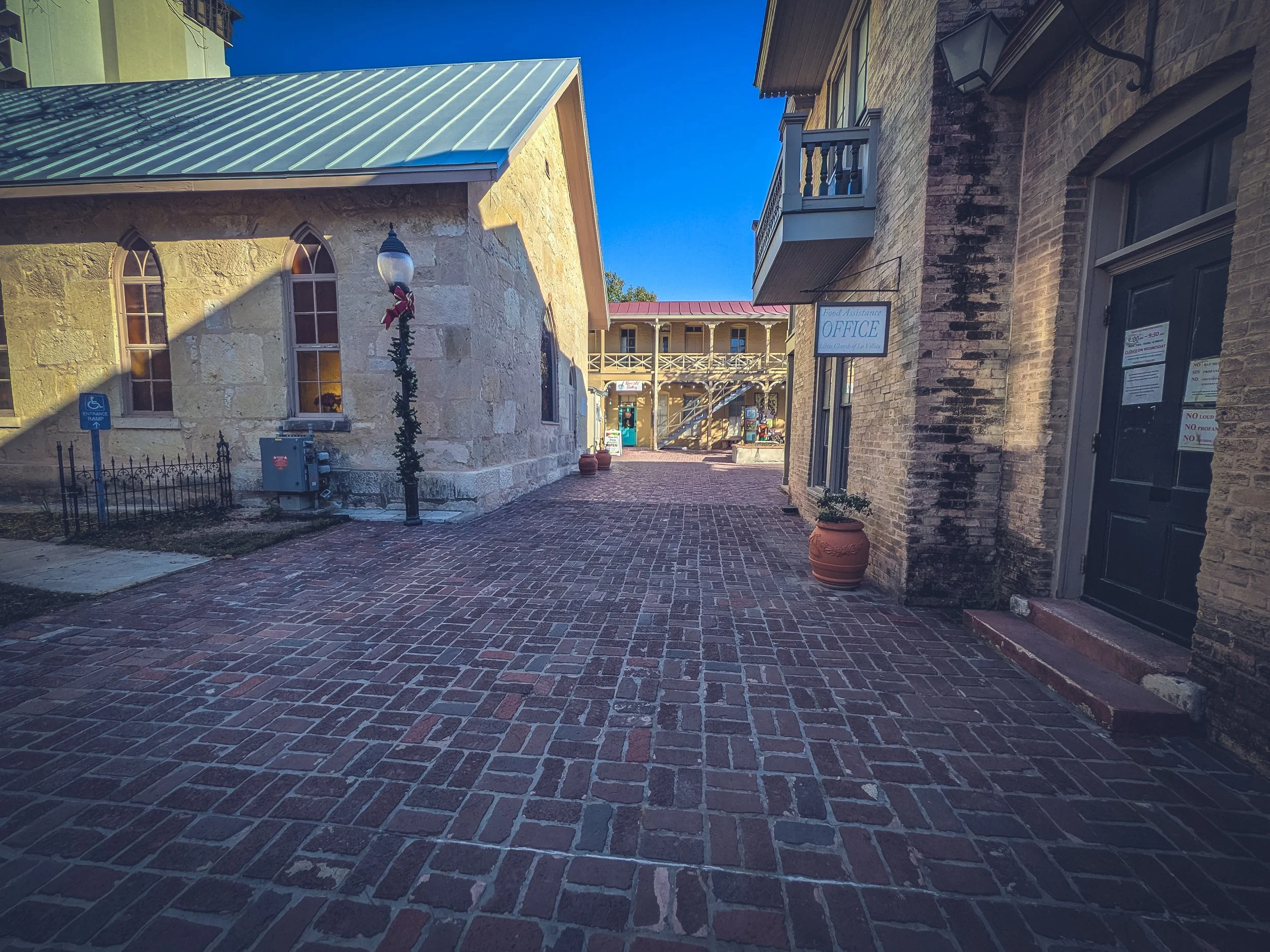 Brick sidewalk in a small town with old stone and brick buildings, a street lamp decorated for Christmas, and potted plants, under a clear blue sky.