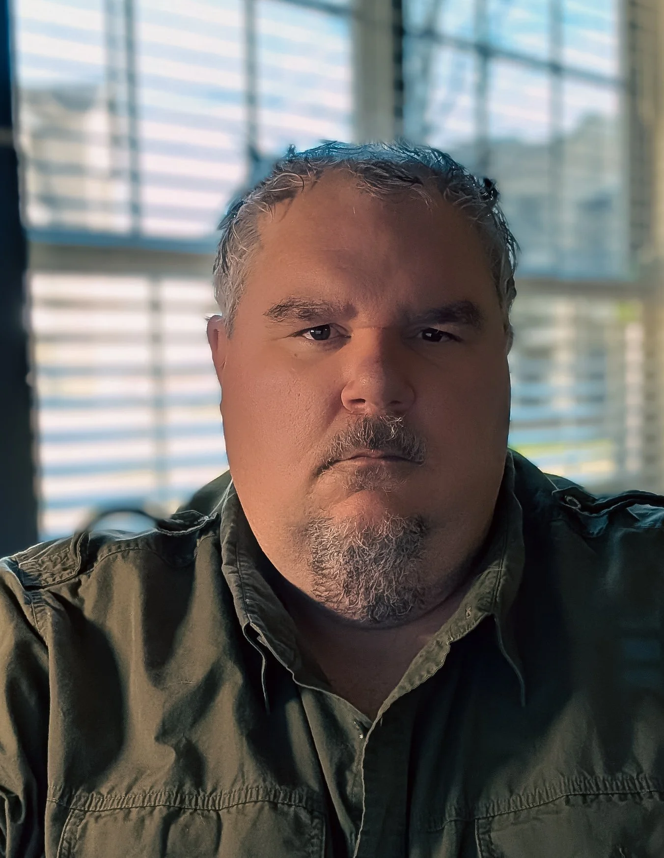 A middle-aged man with gray hair, a beard, and a mustache wearing a dark green shirt, sitting indoors in front of large windows with blinds, looking directly at the camera.