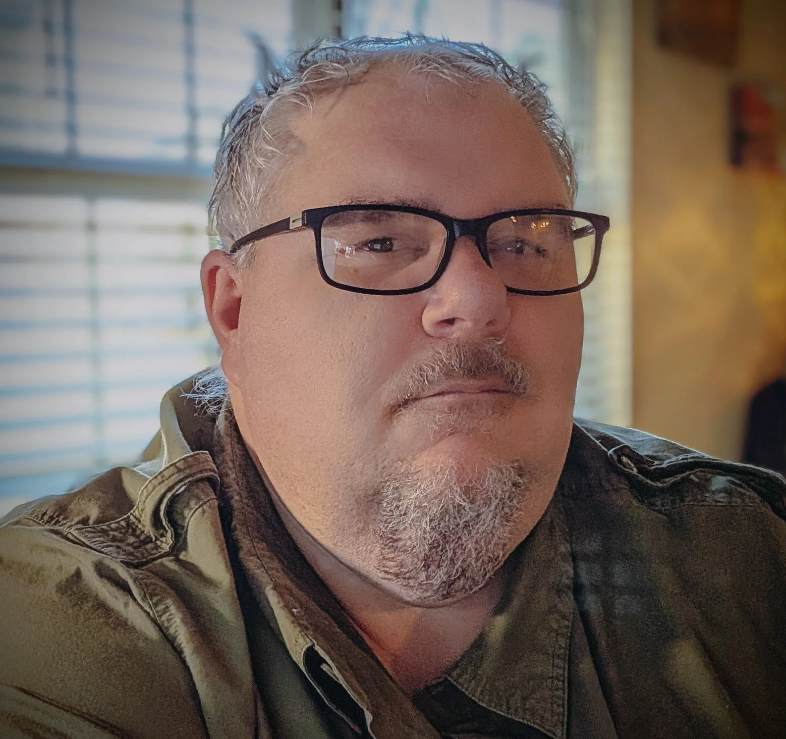 Close-up of a middle-aged man with glasses, gray hair, and a goatee, wearing a military-style shirt inside a room with windows and blinds.