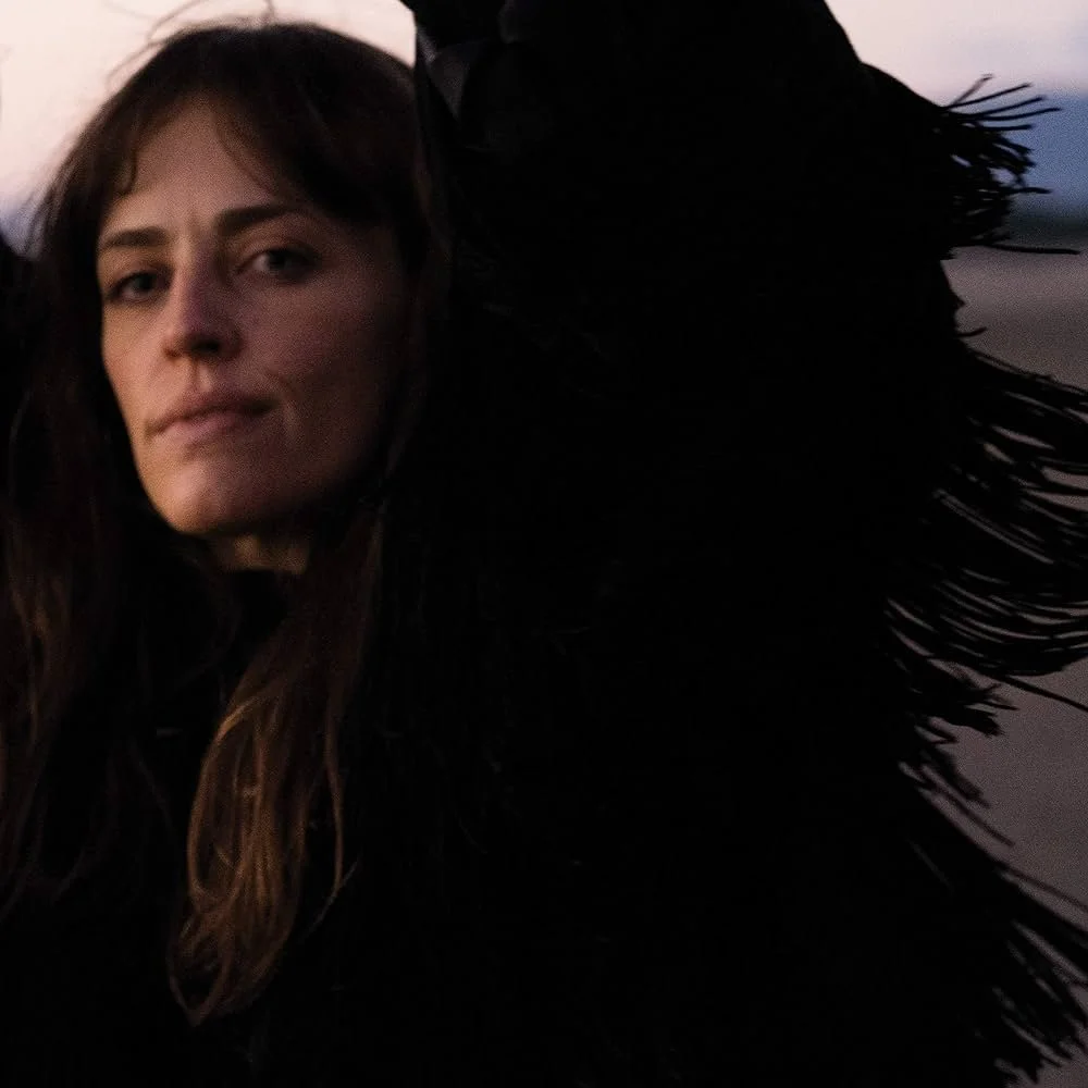 A woman with long brown hair and a neutral expression, wearing a black feathered garment, poses for a selfie against a background of a darkening sky at sunset.