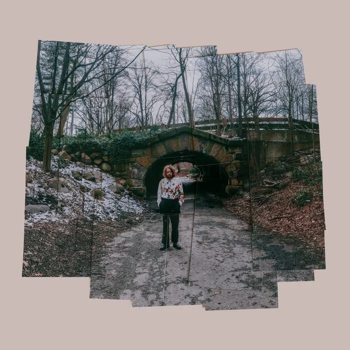 A man standing on a dirt path in front of a stone arch bridge in a wooded area during winter.