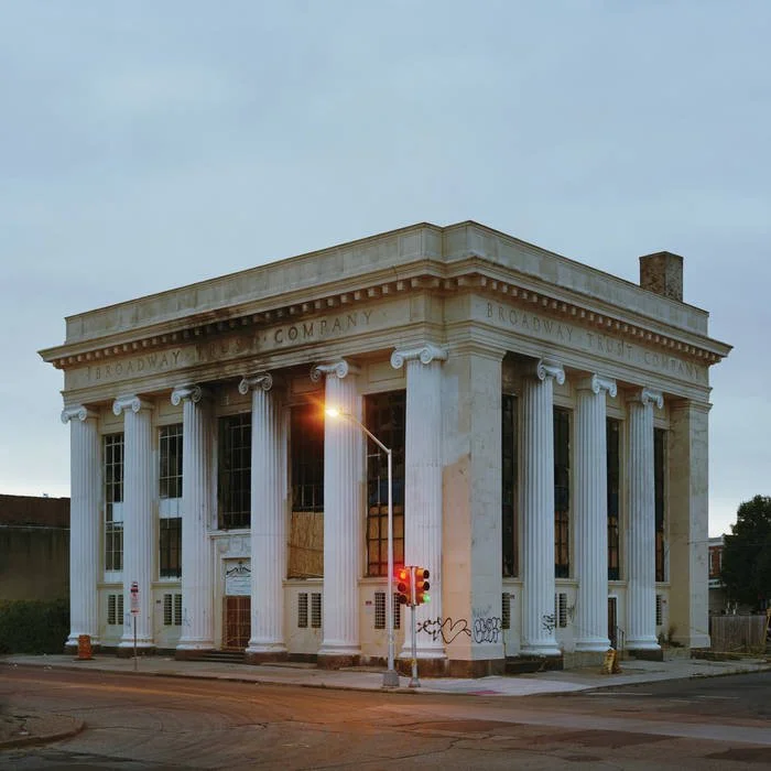 A historic white building with large columns and an inscription reading "Broadway Trust Company" on the frieze, located on a street corner with a traffic light and graffiti on the lower wall.