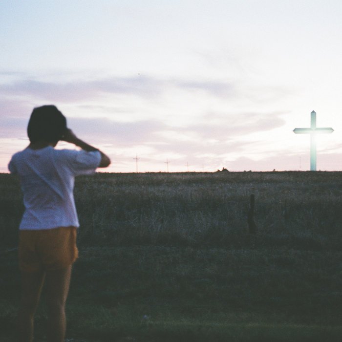 A child with black hair, wearing a white shirt and orange shorts, looking at a distant cross on a hill during sunset.