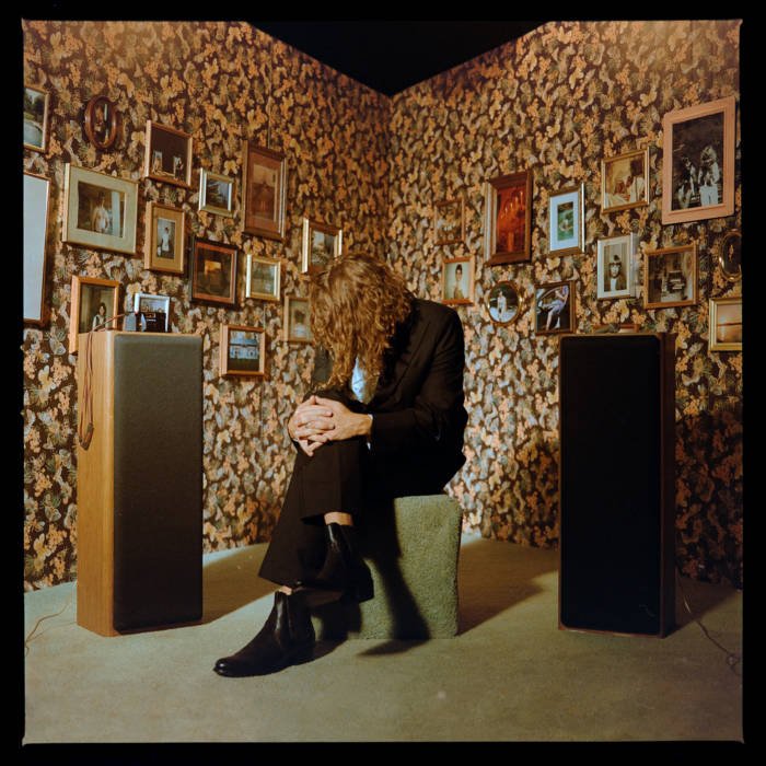 Woman with curly hair sitting on a stool in a room with floral patterned wallpaper and many picture frames on the wall, two large speakers on either side.