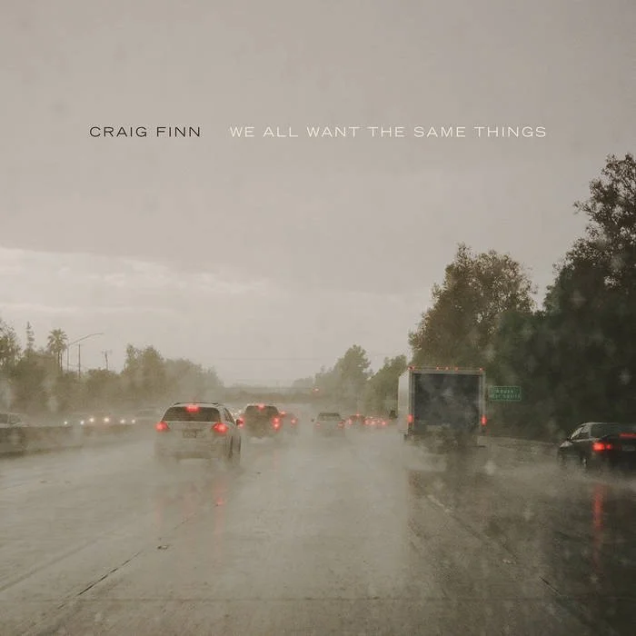 Rainy highway with multiple cars and trucks, trees on the side, overcast sky, album title 'We All Want The Same Things' by Craig Finn