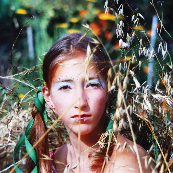 A girl with braided hair and green ribbons, wearing makeup with painted white eyebrows and dots, surrounded by tall dry grass in a natural outdoor setting during daytime.