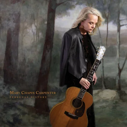 Mary Chapin Carpenter holding an acoustic guitar, standing outdoors with trees and a nature background, wearing a black leather jacket and looking down.