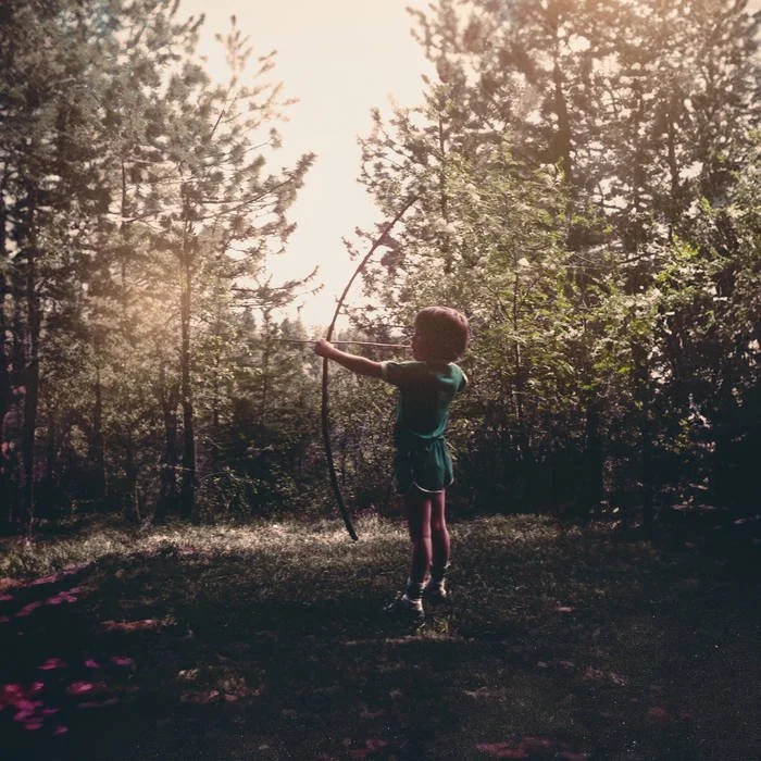 A young child practicing archery outdoors in a forest clearing during daytime.