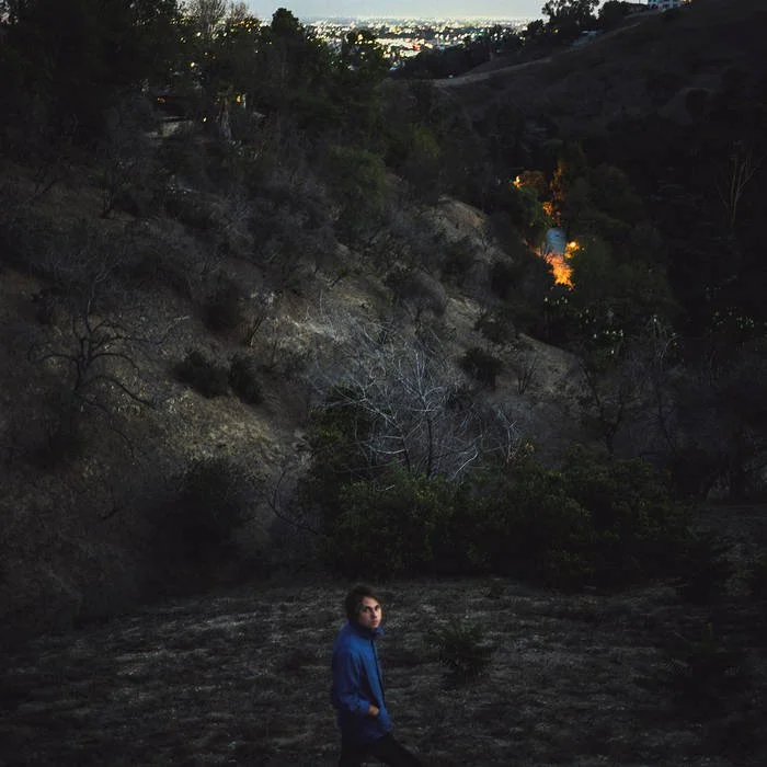 A person stands in a dark, rocky outdoor area with dry, sparse trees and shrubs, overlooking a hillside with distant city lights and a sky transitioning from dusk to night.