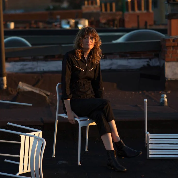A woman with wavy brown hair, wearing a black jacket and black boots, is sitting on a white chair on a rooftop during sunset.