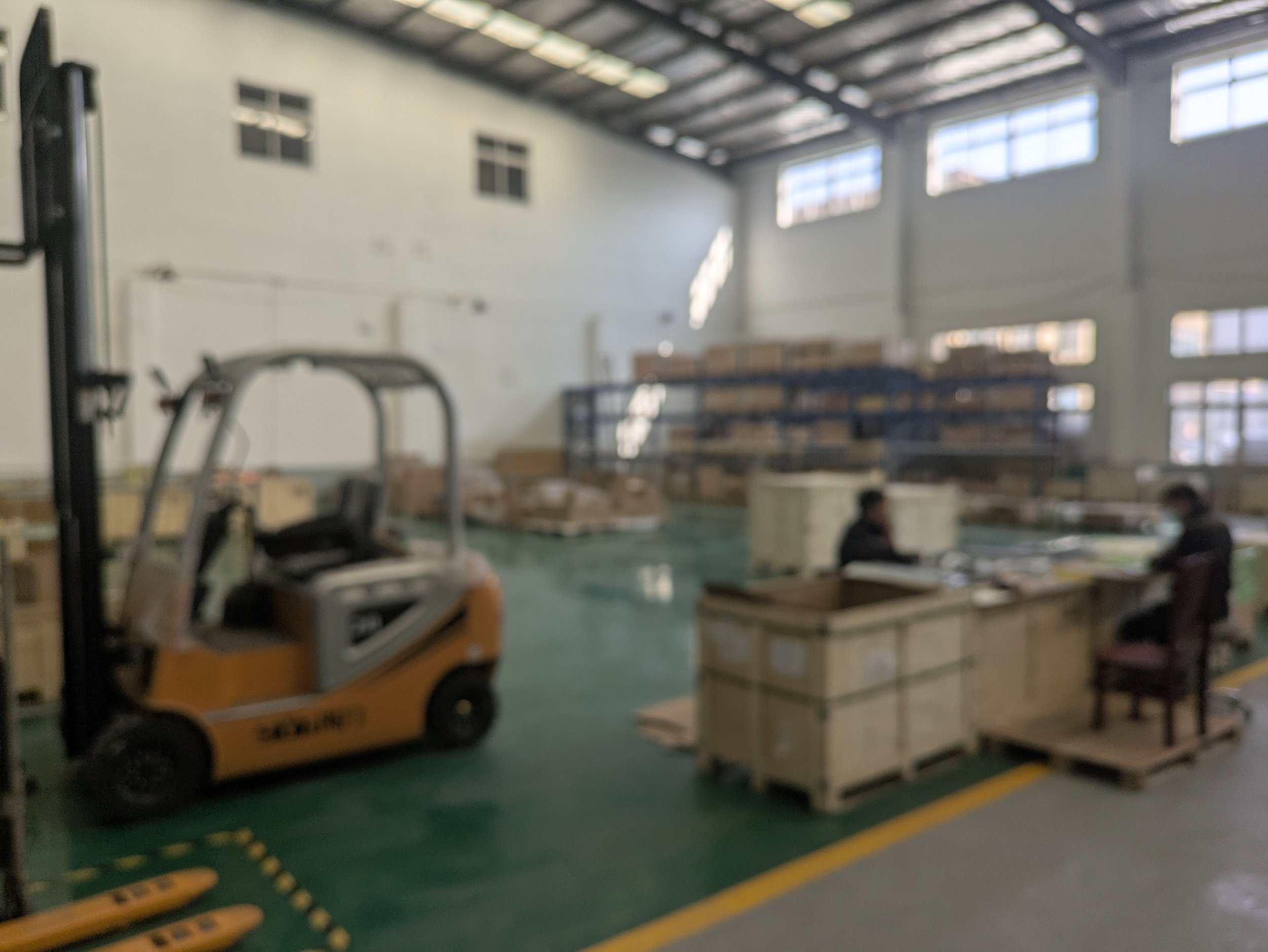 Inside a warehouse with a forklift, wooden crates, and two workers at desks.