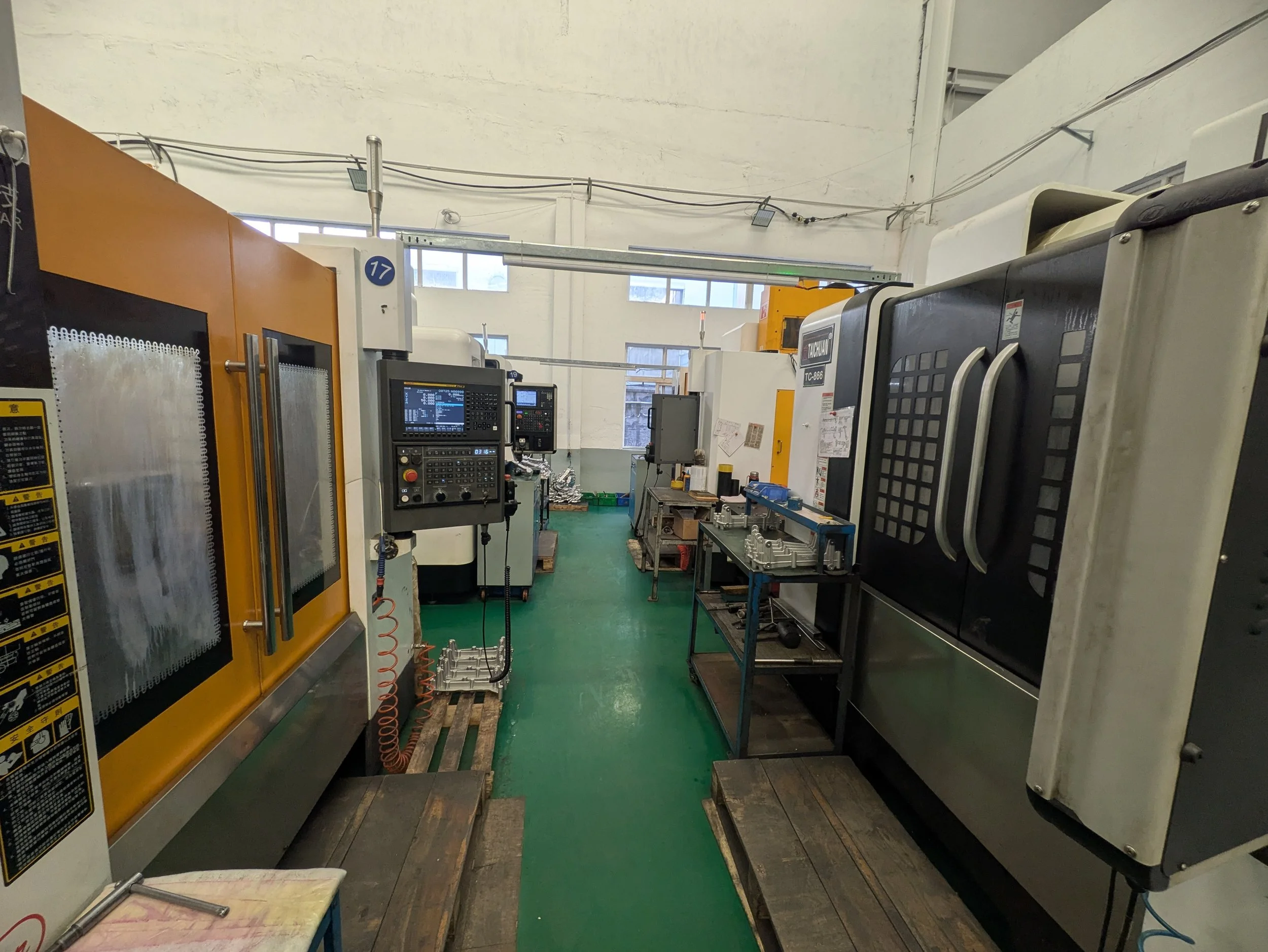 View of an industrial workshop with various machinery, control panels, and workbenches. The floor is green, and there are large windows on the wall allowing natural light inside.