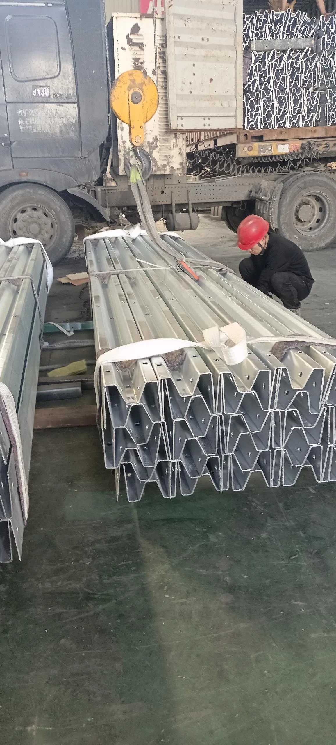 A worker wearing a red safety helmet and black clothing is crouching next to metal structural components on the floor. The components are stacked and secured with white straps, and there is a large truck with a crane hook overhead, used for lifting h