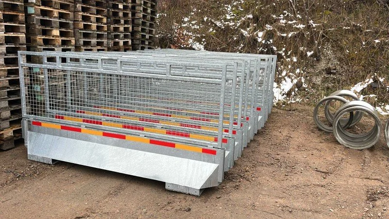 Stacked metal safety barriers and coiled metal pipes on dirt ground outside, with snow on a hillside in the background.