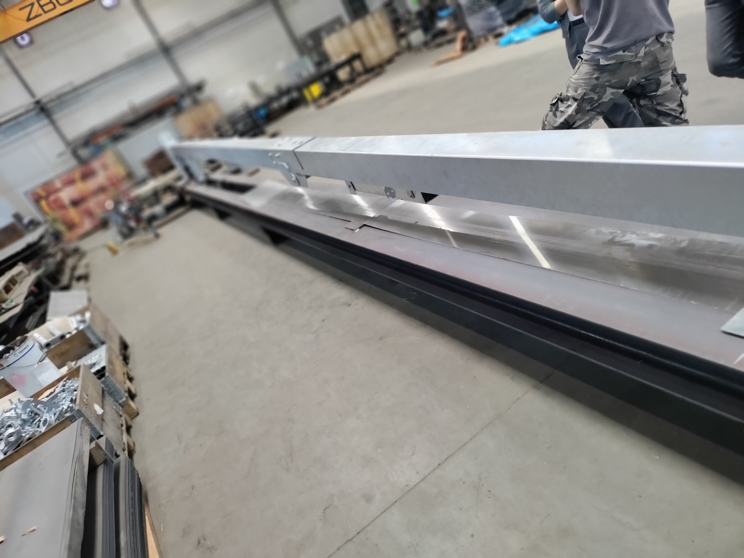 A long metal structure in a workshop or warehouse with various tools and materials around. Partially visible people are standing nearby.
