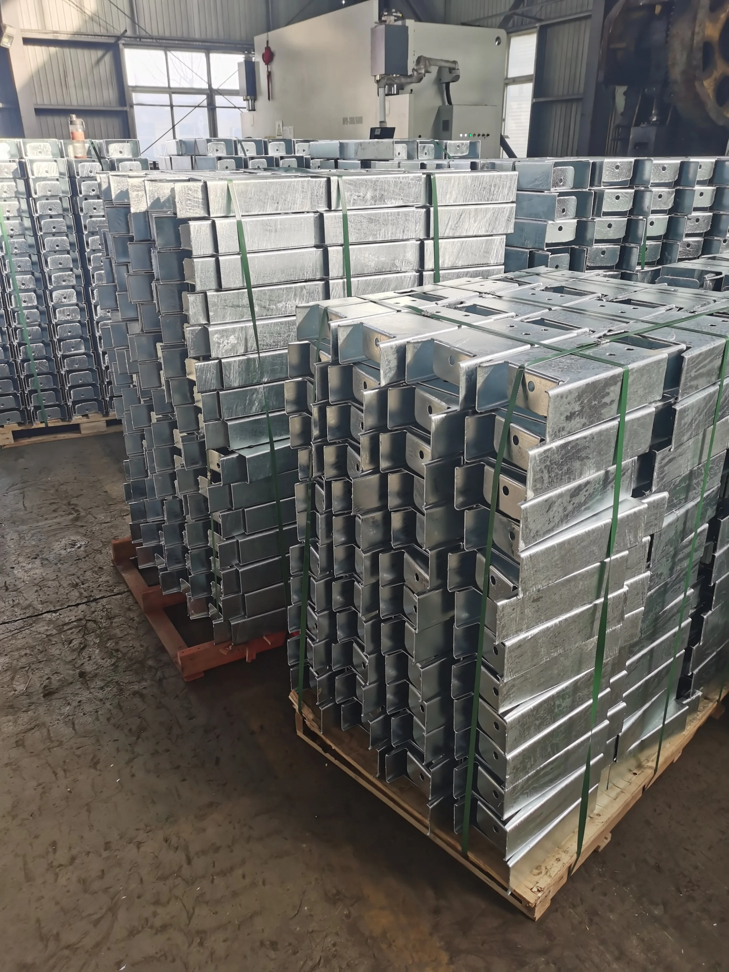 Stacks of metal U-shaped channels secured with green straps on wooden pallets inside a factory or warehouse.