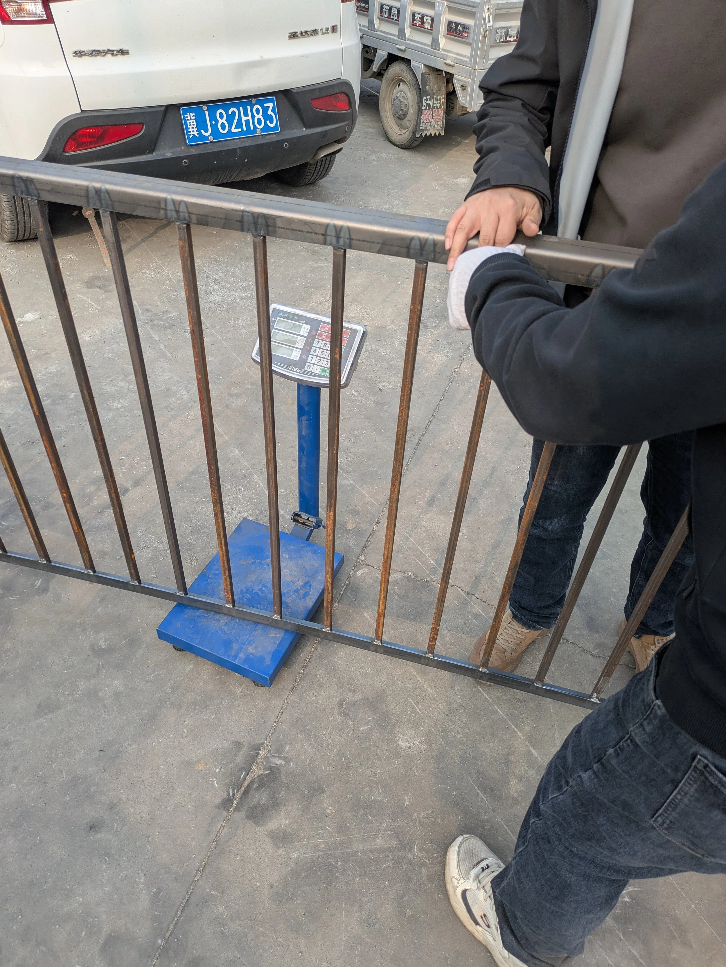 A person in a black jacket is standing behind a metal barricade, holding it with both hands. There is a digital weighing scale underneath the barricade on the ground, and a vehicle with a blue license plate is visible in the background.