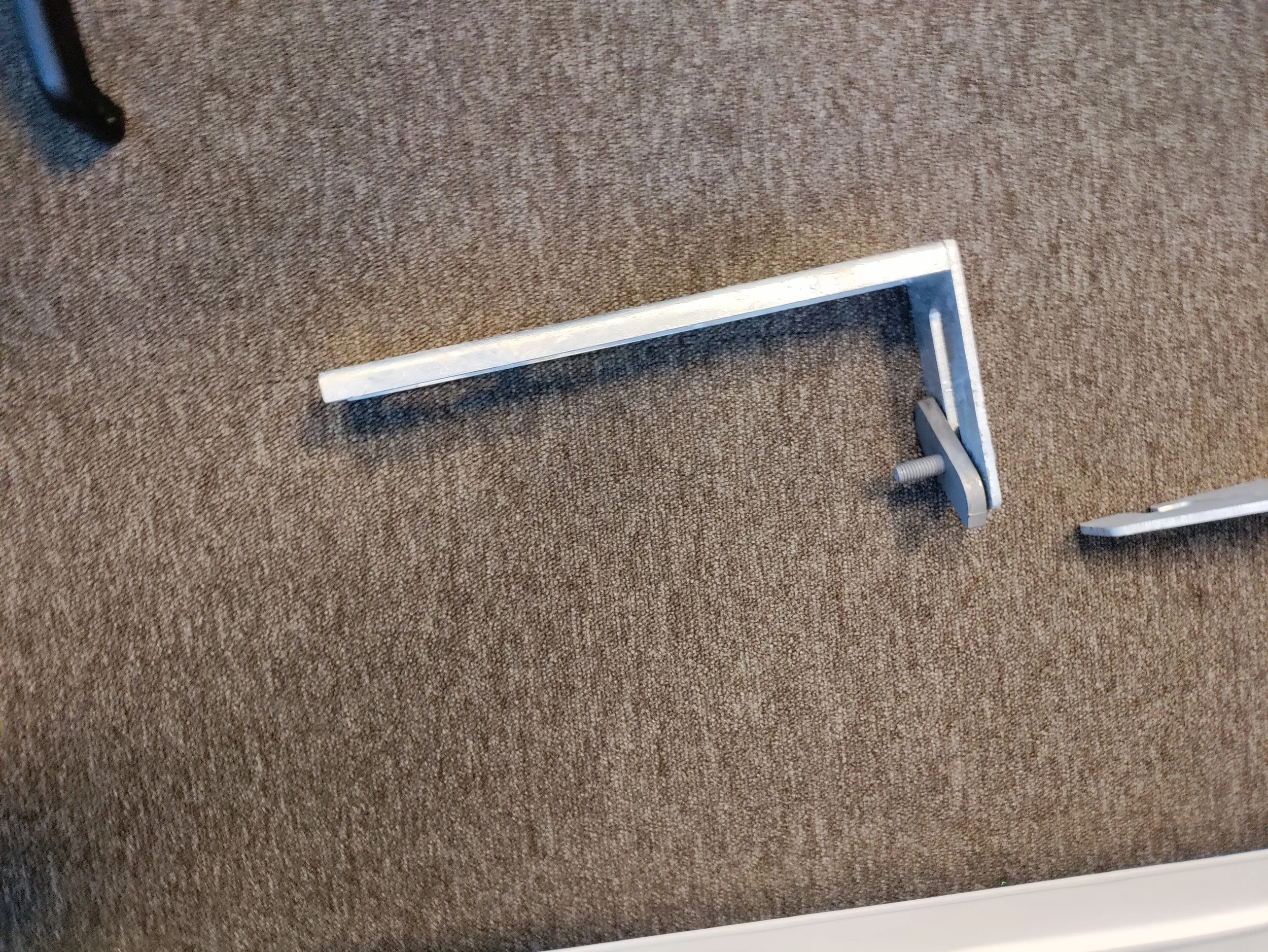 A metal L-shaped furniture leg with a threaded bottom end rests on a brown textured carpet. Part of a black piece of furniture is visible in the upper left corner.