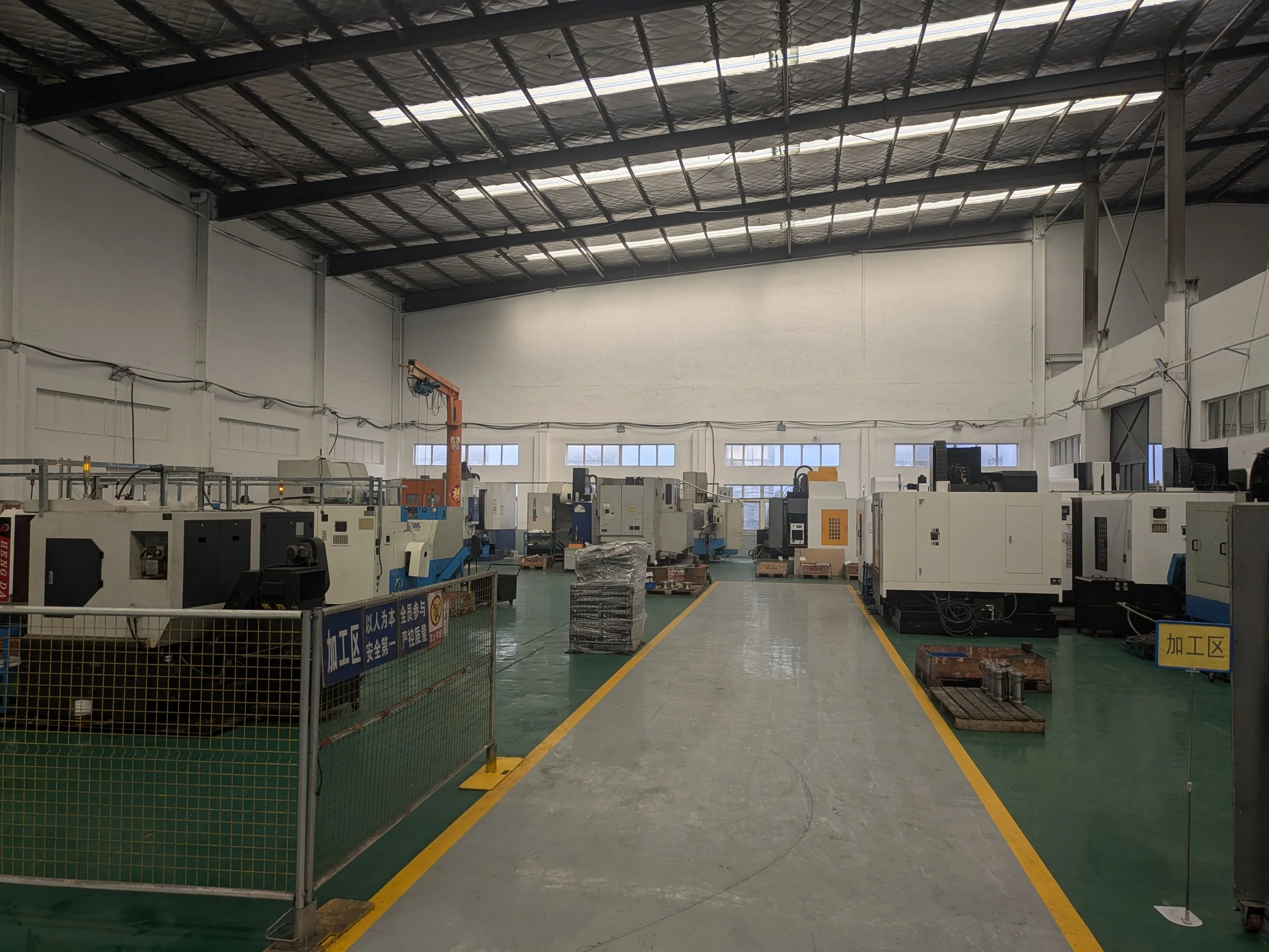 An industrial factory interior with multiple manufacturing machines lined up, a pathway in the center, and safety signs on the fencing.
