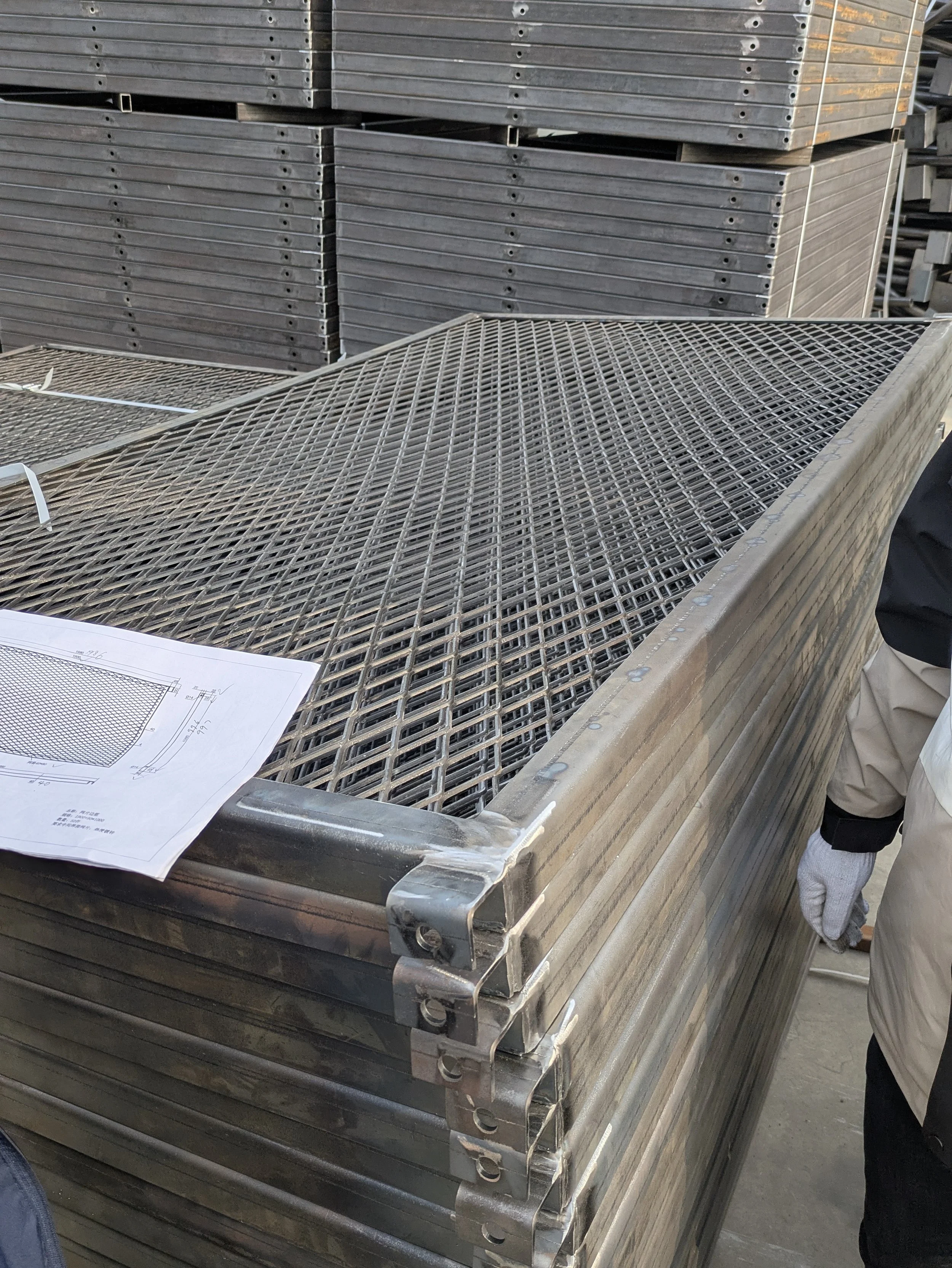 Stacks of metal grates and frames at a construction or manufacturing site, with a blueprint placed on top of one of the stacks.