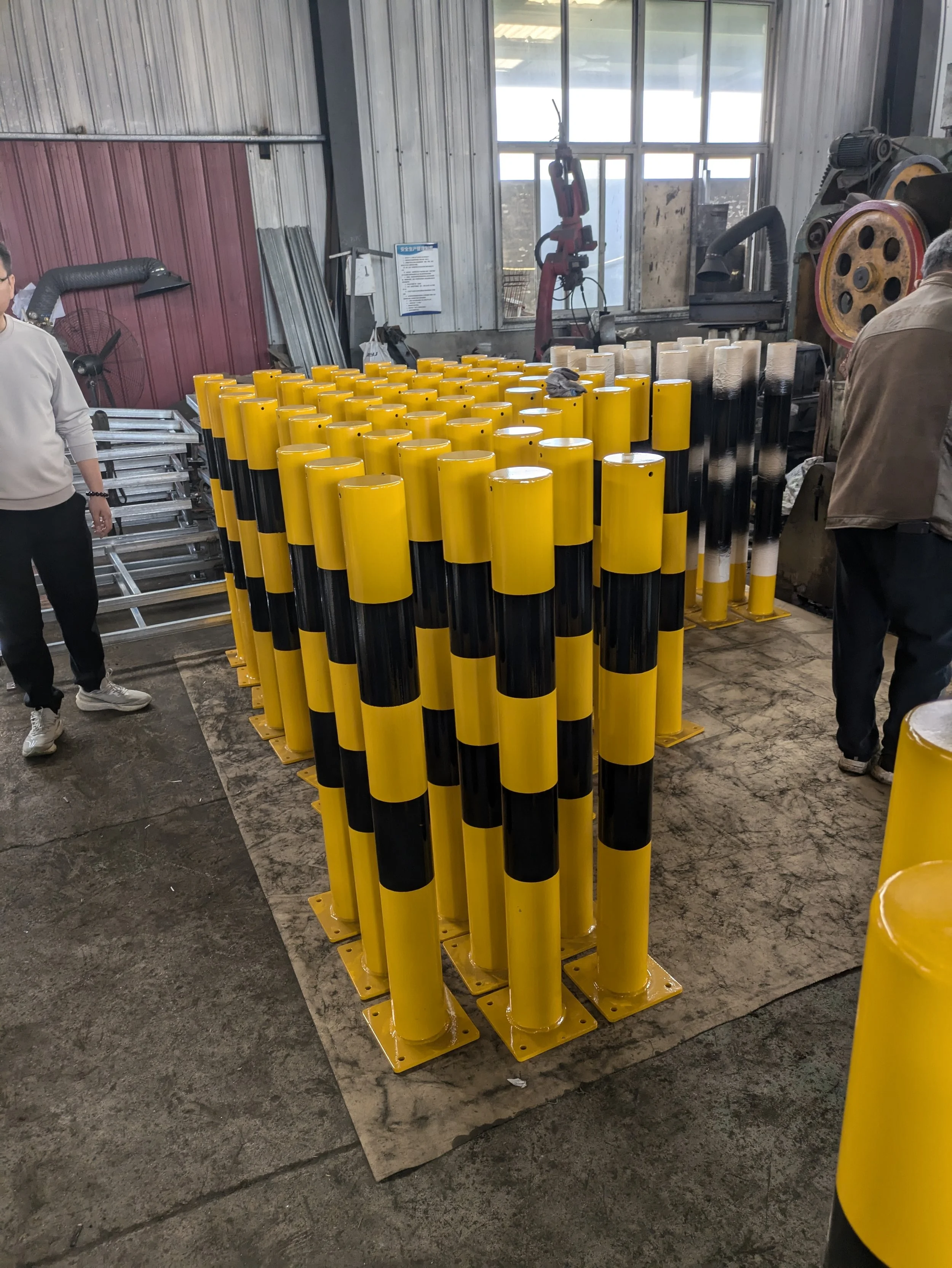 Several yellow and black striped safety barrier posts in a workshop or industrial setting with equipment and people in the background.