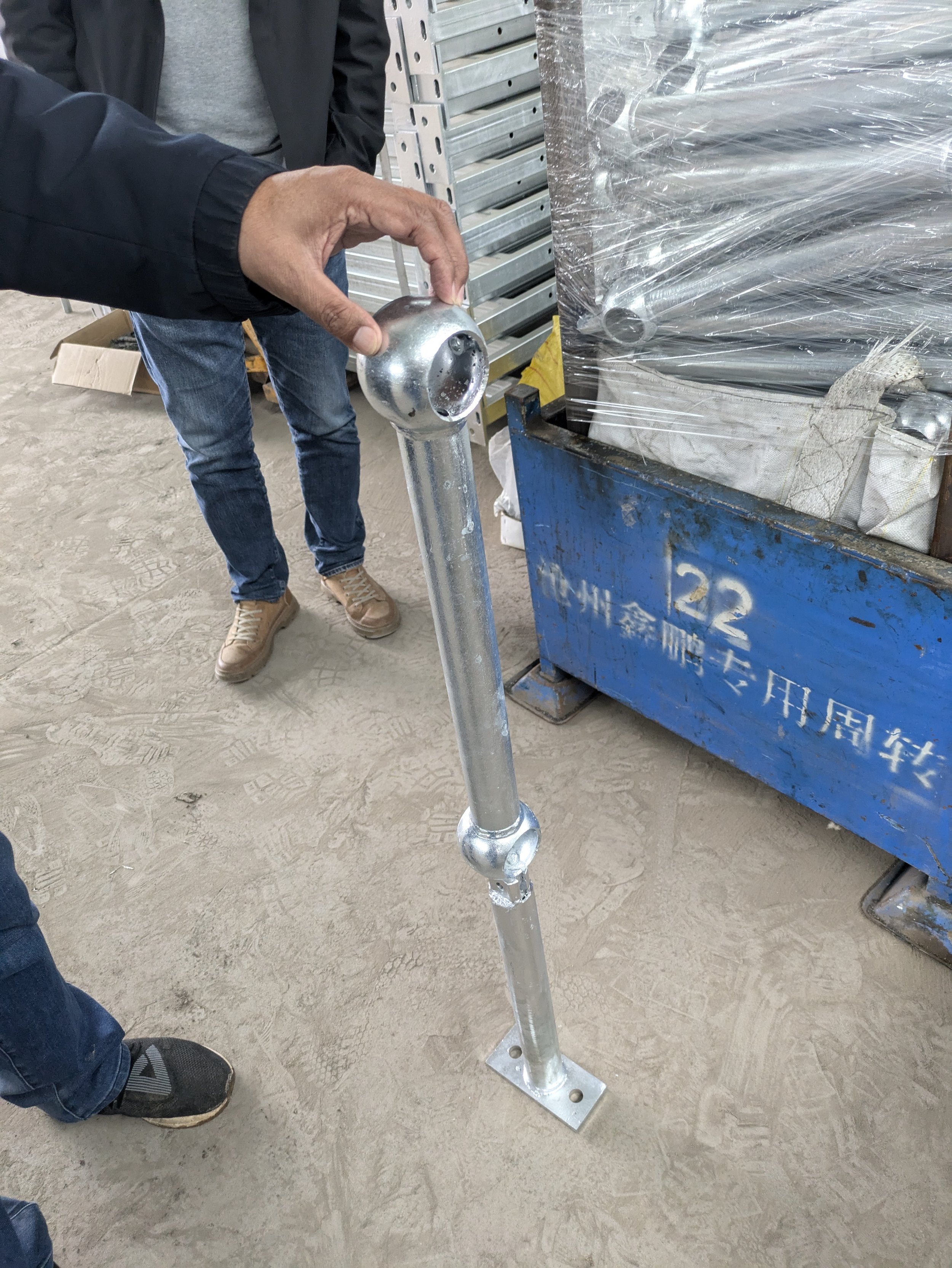 A person holding a metallic handrail post with a spherical top in an industrial or construction setting, with other metal items and equipment in the background.